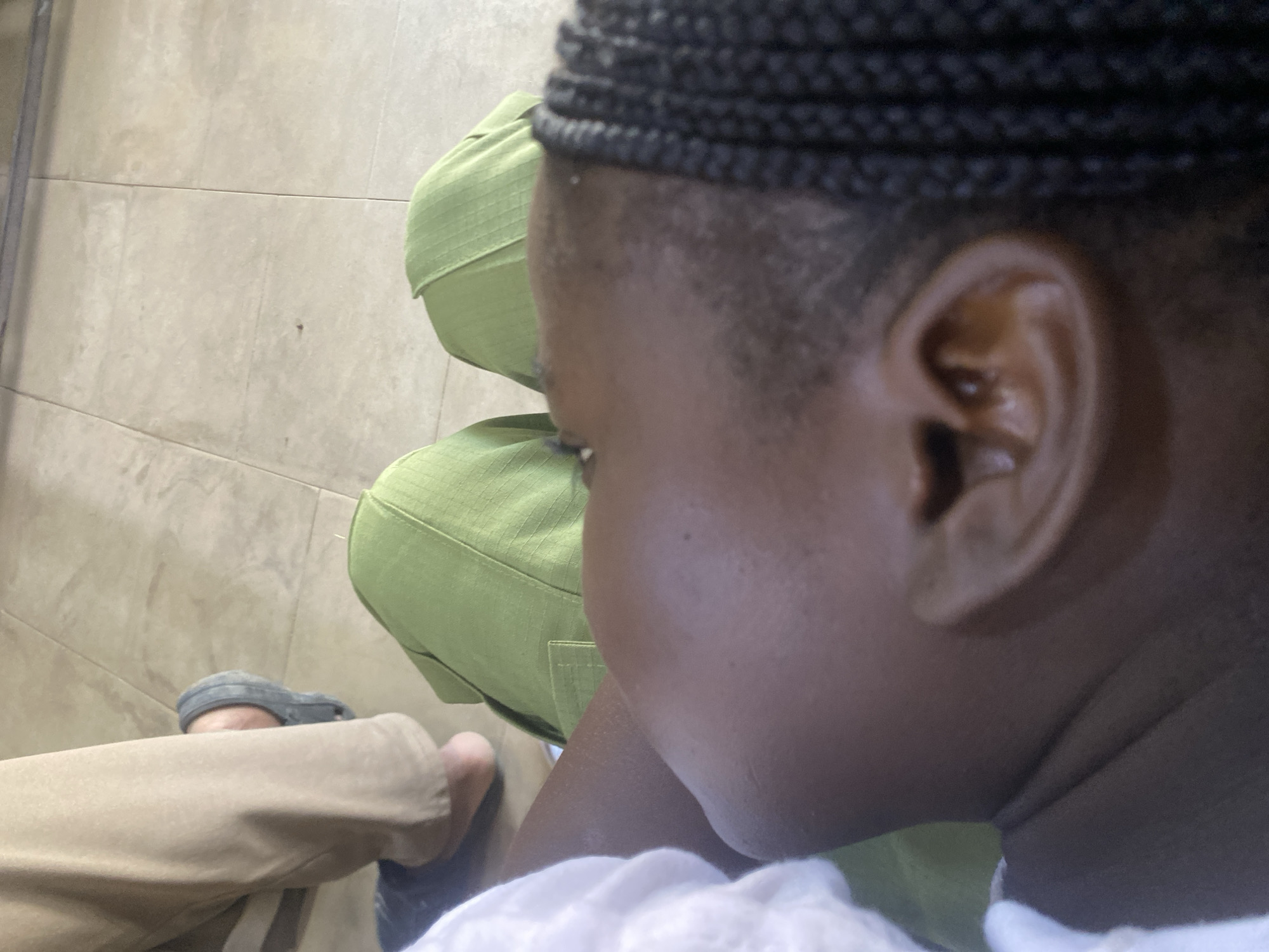 The image shows a close-up of a person's head and upper body, with a focus on their profile and ear. They appear to be wearing a white garment on their left side and green trousers. In the background, another person's legs are visible, wearing khaki trousers and grey clogs. The floor is tiled with large, light-colored square tiles. The scene is shot from a high angle, looking down at the person. It is impossible to determine the specific location in Jimeta, Nigeria, based solely on this image.