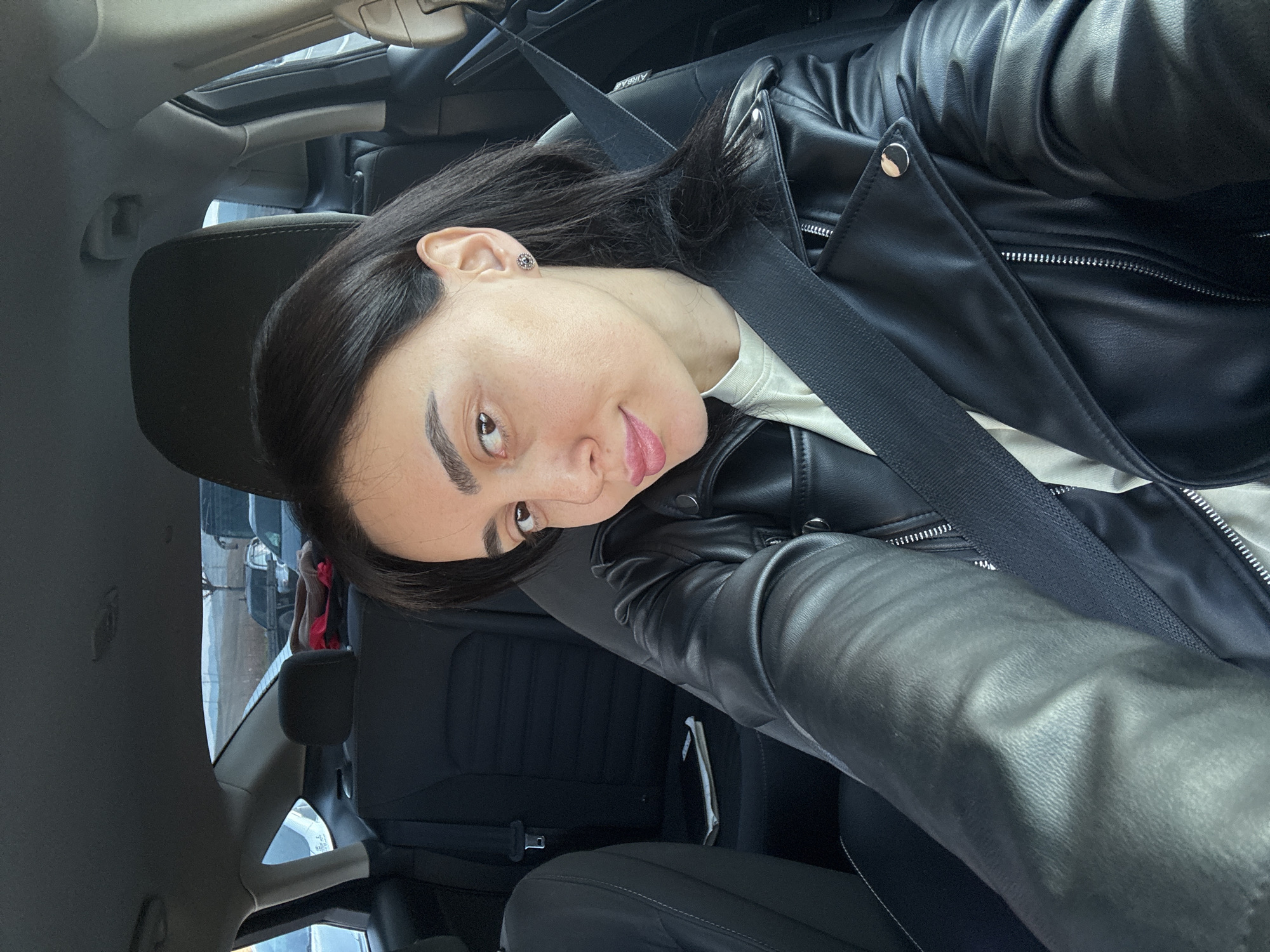 A woman with dark hair and fair skin is sitting in the driver's seat of a car. She is wearing a black leather jacket and a white top, and a seatbelt is fastened across her chest. The car's interior is dark, with black seats and dashboard. The car has a sunroof.
The location appears to be a street in Tbilisi, Georgia, based on the blurred view through the car windows showing other vehicles and buildings.