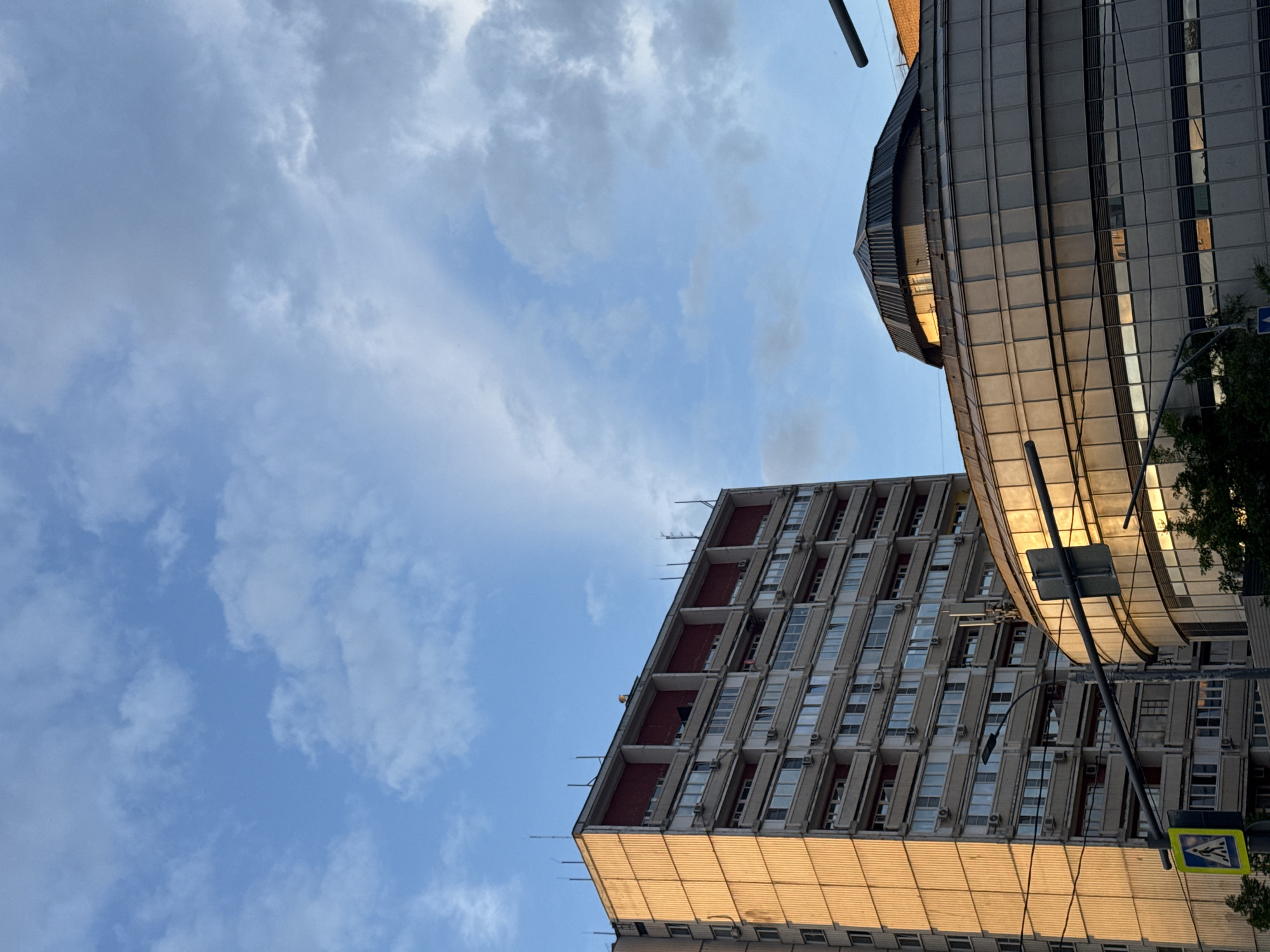 The image captures an urban landscape featuring two prominent buildings under a partly cloudy sky, viewed from a low angle looking upwards. The scene is illuminated by the warm, golden light of the late afternoon or early evening sun.
On the left, a tall, multi-story building displays a rectilinear architectural style, characterized by a repetitive pattern of windows, balconies, and external air conditioning units. Its lower facade, or an adjacent structure, reflects the golden light vividly, with bird deterrent spikes visible along its edges.
To the right, a more contemporary building features a curved, glass-clad facade with a distinct, angular dark roof structure. This building also reflects the warm light, creating bright golden reflections on its surfaces.
In the bottom right corner, partially obscured by the modern building and some tree foliage, a dark pole supports a yellow and blue pedestrian crossing sign, which depicts a person walking. The sky above is a mix of clear blue and diffuse white and grey clouds. The overall impression is one of a busy urban environment, with contrasting architectural styles under a serene sky. No people are visible.