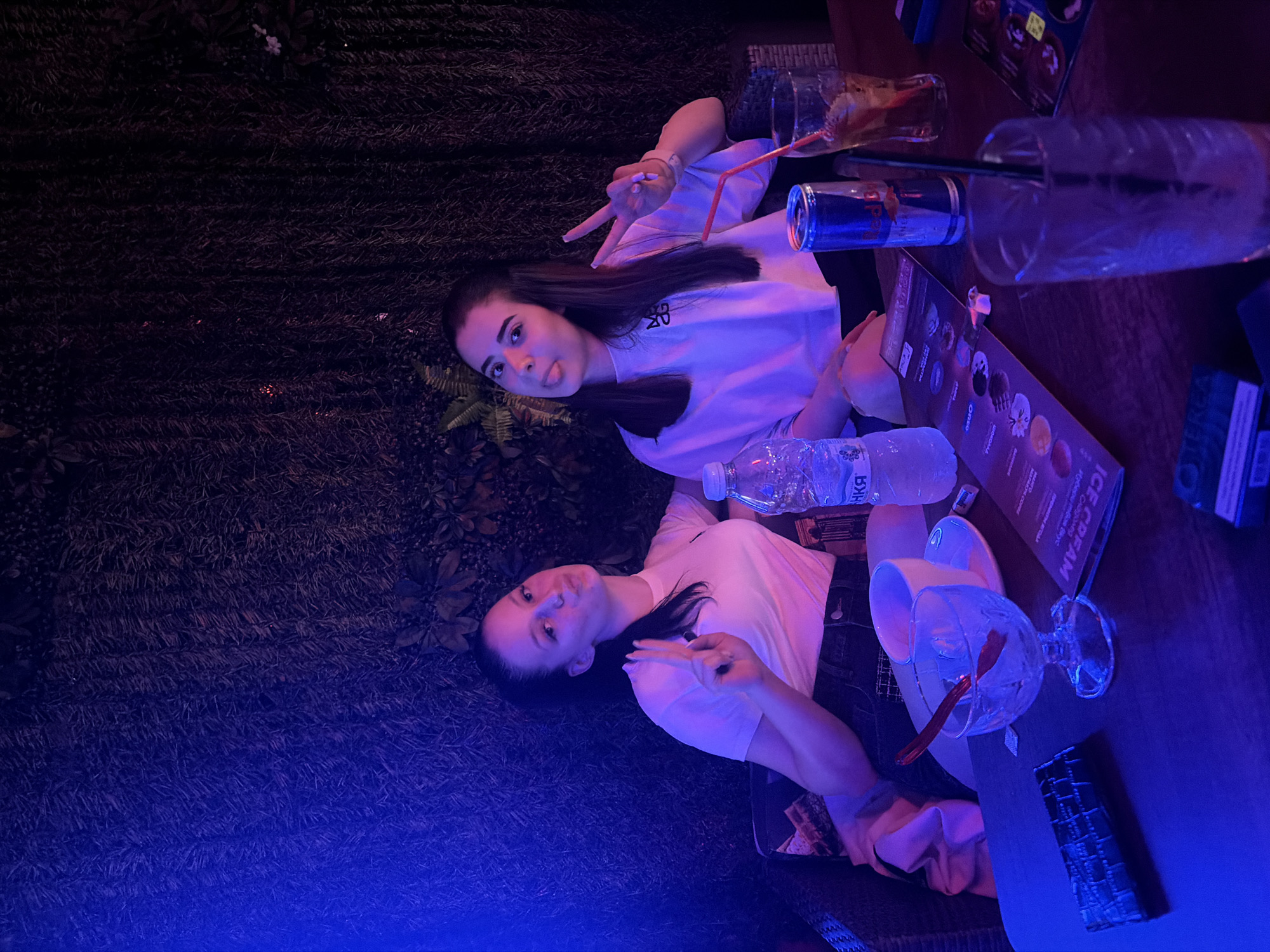 Two young adult females are seated at a dark-colored table in an establishment located in Elhovo, Bulgaria, characterized by dim, artificial blue and purple lighting and a background wall covered in dense, dark green foliage.
The female on the left has dark hair pulled back, wears a white t-shirt and dark bottoms, and is shown with puckered lips and her right hand displaying a two-finger peace sign. The female on the right has long dark hair, wears a white t-shirt with a dark logo, and is sticking out her tongue while her left hand displays a two-finger peace sign.
The table in front of them holds various items: a dark rectangular object (possibly a wallet or phone case), a clear glass dessert dish with a red spoon, a white ceramic teacup and saucer, a clear plastic water bottle with a partially visible Cyrillic label, a menu featuring "ICE CREAM" and multiple options with circular images, a silver and red Red Bull can, a tall glass containing a beverage with ice, fruit, and a red straw, and another tall, empty glass with a black straw positioned in the foreground. The seating is dark and appears cushioned.