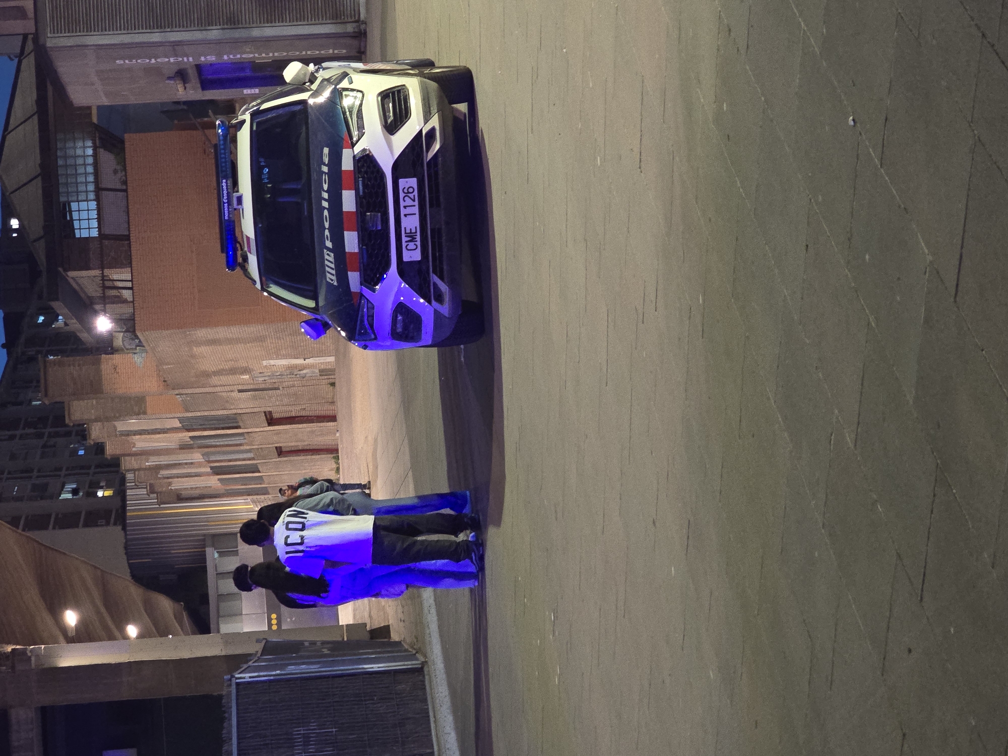 The image depicts a nighttime urban scene in Cornellà De Llobregat, Spain, featuring a police vehicle and several individuals on a paved street or square.
In the foreground, a white and dark blue police SUV is parked, with its blue lights illuminated. The side of the vehicle clearly displays "Policia" and the red and yellow stripes of the Catalan flag, indicating its affiliation with the regional police force. The front license plate is visible with the alphanumeric code "CME 1126," and the light bar on the roof bears the text "mossos d'esquadra," the official name of the Catalan police.
Adjacent to the police car, three individuals are standing on the dark-colored paved ground. Their faces are not discernible due to the lighting and their orientation. One person wears a white long-sleeved shirt with the word "ICON" printed in black on the back, strongly illuminated by the blue light from the police vehicle. This blue light casts a distinct glow on their lower body and the surrounding pavement.
In the background, various buildings with brick and concrete facades are visible, along with some windows. Part of an overhead structure or building reveals partially visible text that reads "aparcament St Ildefons," which translates to "St Ildefons Parking," providing a specific location cue. The overall dim lighting confirms it is nighttime, with artificial lights scattered throughout the scene.