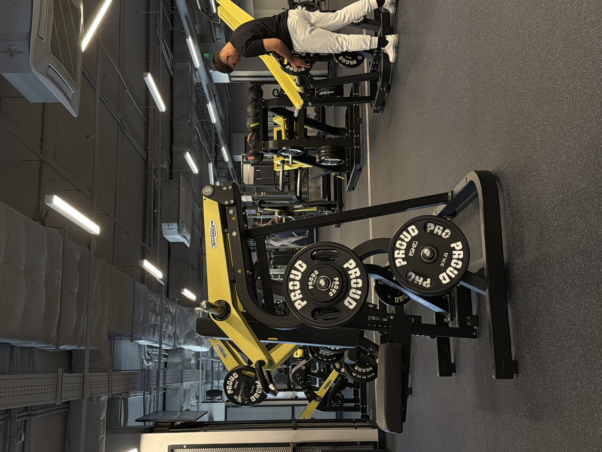 The image shows a gym interior with a person using a lying leg press machine. The machine is mostly black with yellow accents and has weight plates labeled "PROUD 15KG" and "PROUD 33LB". The person is wearing a black shirt, white pants, and white shoes. The gym has a gray floor and the walls and ceiling have visible ductwork and lighting fixtures. The location is not specified and the city of Plovdiv, Bulgaria, is not confirmed by the given image.