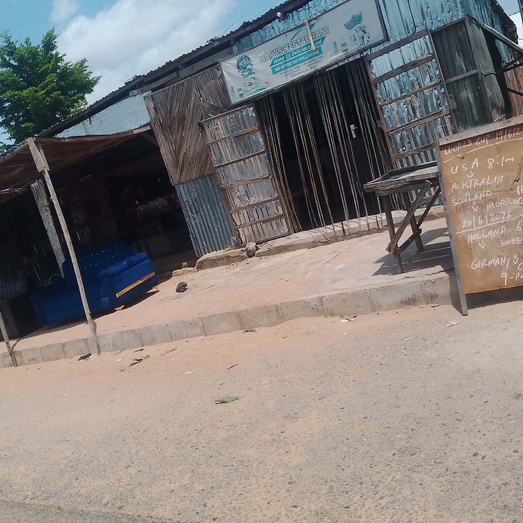This image depicts a street scene in Jalingo, Nigeria. In the foreground, a dirt road is visible with scattered debris. A concrete barrier runs along the side of the road.
Further back, a small building constructed with corrugated metal and wood is the main subject. A banner for "HAPY'S RESTAURANT - Home Of Delicious Food" hangs on the front of the building, advertising "Indomie/Egg, Pounded Yam, Rice Food, Semolina, and Pesc Soup". Adjacent to the restaurant, a wooden easel with a chalkboard displays a schedule for World Cup matches. The chalkboard lists matchups such as "USA 8-1pm", "AUSTRALIA v MORRO", "20/6/2026", "HOLLAND 6", "v SWEE", and "GERMANY 8-9pm".
To the left of the building, a bright blue couch with gold accents is placed on the dirt. In the background, a lush green tree stands tall against a cloudy sky. The overall impression is of a casual, local establishment, possibly in the afternoon given the lighting. The scene suggests a community gathering place, likely for watching the World Cup.