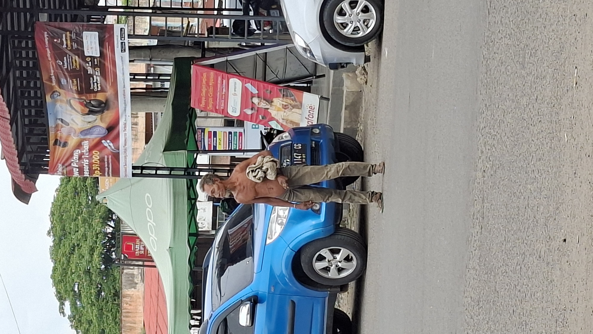 The image depicts an outdoor scene in Lhokseumawe, Indonesia, likely on a street or in front of a commercial area. A man, shirtless and wearing tan trousers and sandals, stands in the middle of the frame, holding a bundle of fabric. To his left, a blue SUV is parked, and to his right, a portion of a silver car is visible. In the background, a green tent with the "OPPO" logo is set up, suggesting a promotional event or retail space. Behind the tent, a red and white banner advertising "Aksesoris Terbaik Rp 39.000" (Best Accessories for Rp 39,000) is displayed. Further in the background, lush green trees are visible, with a building structure partially obscured by the tent and banner. The sky is overcast, indicating a cloudy day. The overall impression is a snapshot of everyday life in an urban setting, with elements of commerce and transportation.