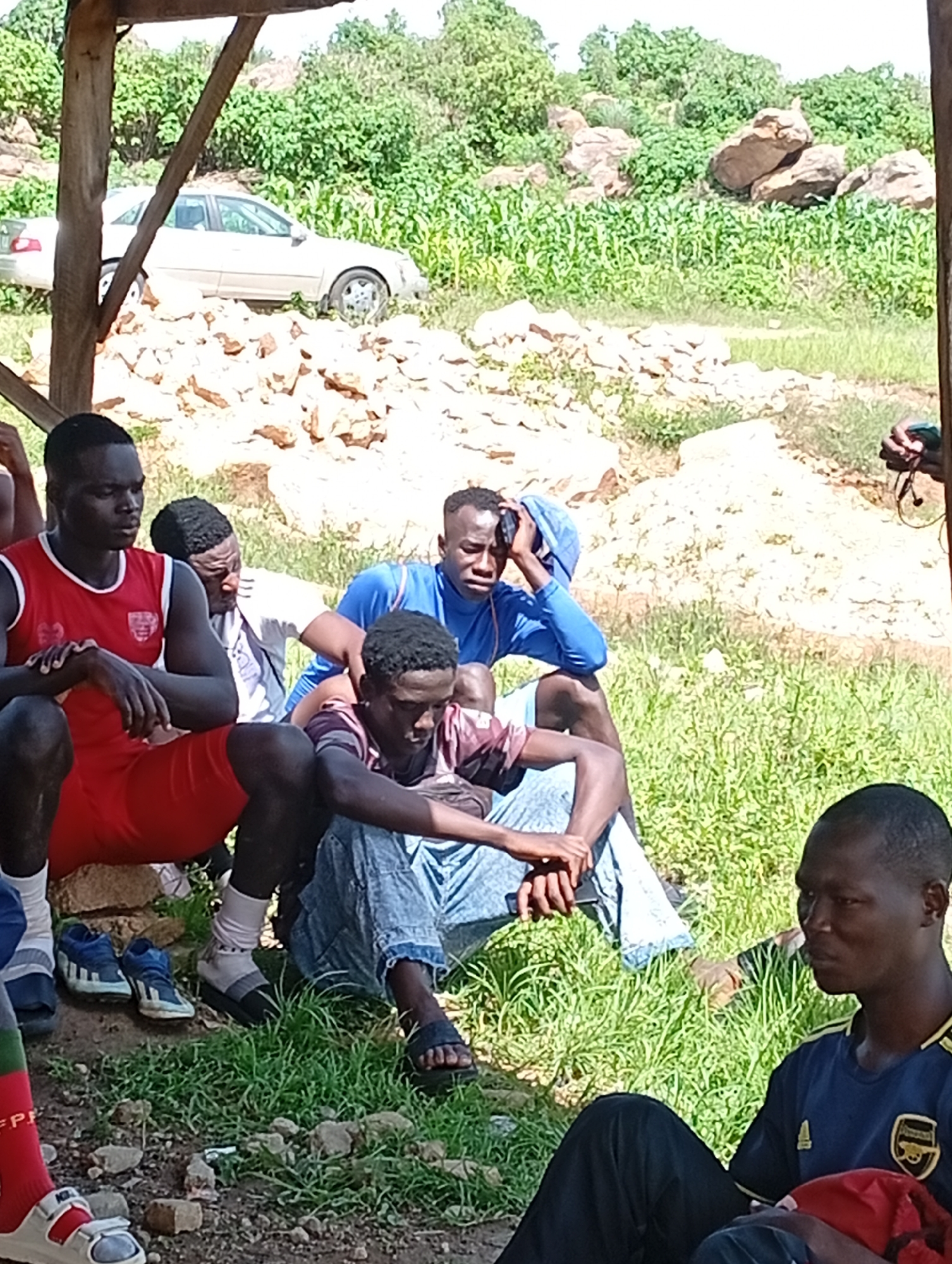 The image depicts a group of young men gathered outdoors, seemingly in a casual setting with a backdrop of lush green vegetation and rocky terrain. The scene appears to be in a semi-rural or informal area within the city of Jos, Nigeria, characterized by the natural landscape and simple overhead shelter.
In the foreground, several individuals are seated or resting. One young man in a red athletic top and shorts is prominently positioned on the left, with his arms crossed. To his right, another man in a blue long-sleeved shirt is looking down. In the center, a man in a patterned shirt and denim jeans is seated, holding a phone. Further to the right, another young man in a dark blue t-shirt with yellow accents and an emblem is visible, looking to his left.
Scattered around the seated individuals are various objects including shoes, socks, and what appears to be a red bag. A light-colored sedan is parked in the background, partially obscured by vegetation and rocks. The environment suggests an outdoor gathering, possibly after a sporting activity or a break in a day's activities. The presence of the car and the general appearance of the surroundings do not definitively place the location in a specific urban district of Jos, but it is consistent with areas that blend natural landscapes with residential or community spaces.