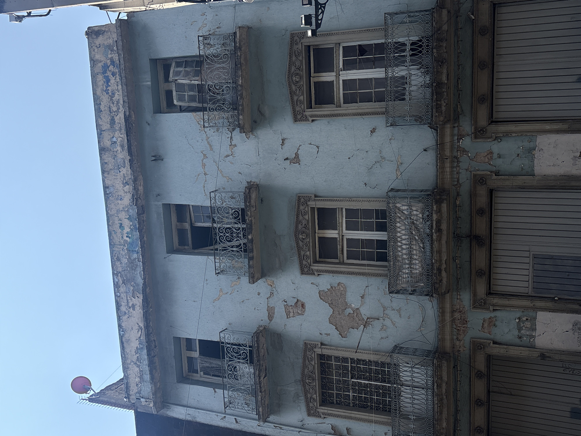 The image shows a dilapidated building in Mexico City, Mexico. The building has three stories, each with windows. Some windows have decorative iron bars, and some have wooden shutters. The building's facade is peeling and cracked, exposing the underlying layers of paint. The sky is visible in the upper left corner. A red object resembling a satellite dish is attached to the roof.