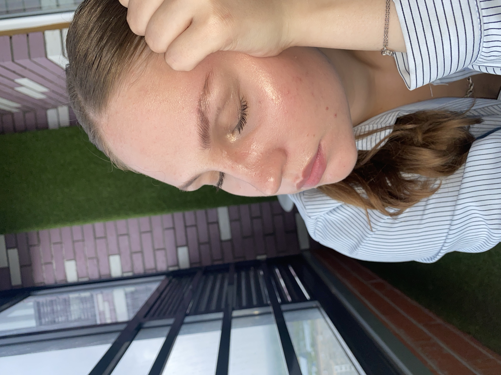 A young woman with light brown hair is leaning against a window, her hand resting on her forehead. She is wearing a blue and white striped shirt. Her eyes are closed and her face is tilted downwards. The background shows a window with a view of buildings and a brick wall with a green vertical element. This appears to be a balcony or outdoor space overlooking a cityscape, possibly in Izhevsk, Russia, given the context.