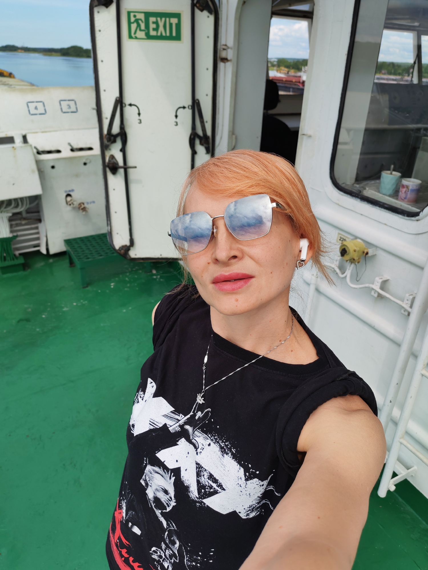 A woman with orange hair and sunglasses is taking a selfie on the deck of a ship. She is wearing a black t-shirt with a graphic print and a silver necklace. She has a white earbud in her left ear. The deck is painted green, and there is a white railing and a white structure in the background. A green sign with an "EXIT" symbol is visible on a white door. The ship appears to be docked, with water and land visible in the distance.