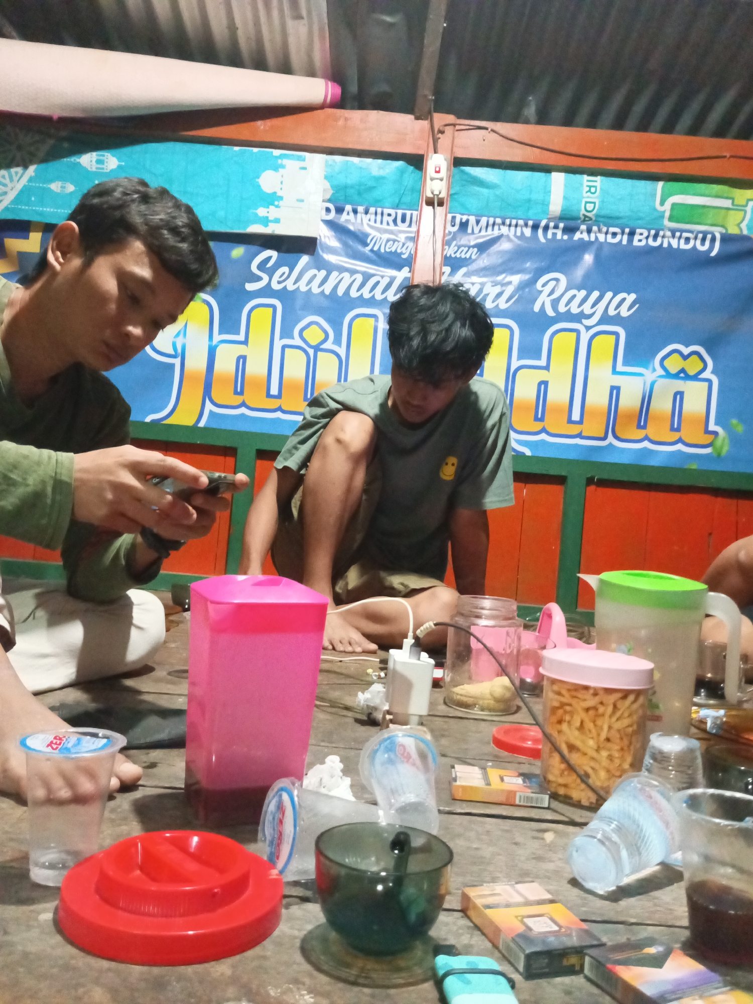 In a dimly lit, outdoor setting, likely in Tumampua, Indonesia, two young men are the primary focus. One man, wearing a green t-shirt, is seated and looking down at his phone, which he holds in his hands. He wears a bracelet on his left wrist. Next to him, another young man, also in a green t-shirt with a yellow smiley face graphic, is crouched down, looking towards the ground.
The scene is set on a rustic wooden table littered with various items. A large pink plastic container dominates the left side of the table. To its right, a clear glass jar contains a yellowish substance. In the center, a pink-lidded jar is filled with what appears to be fried snacks. A green pitcher with a white lid is positioned towards the right. Several plastic cups, some empty and some containing liquid, are scattered around the table, along with a dark-colored bowl and a red plastic lid. Two packs of cigarettes are also visible.
Behind the men, a blue banner with white text and stylized depictions of lanterns and mosques serves as a backdrop, suggesting a celebratory or festive occasion. The overall lighting is dim, with some artificial light sources illuminating the scene. The environment appears to be an informal gathering or hangout spot.