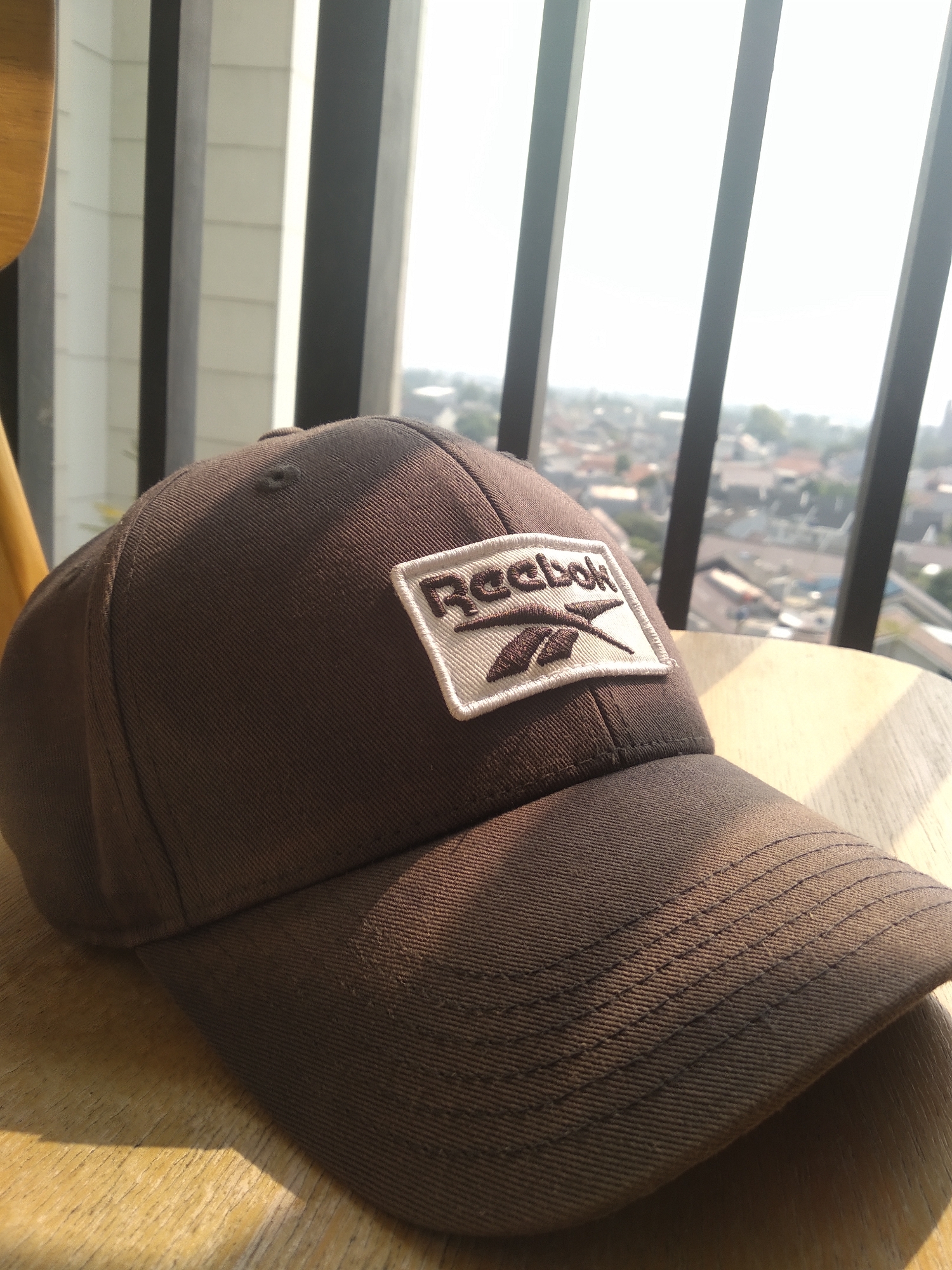 A close-up, eye-level shot shows a brown Reebok baseball cap with a white patch featuring the Reebok logo, lying on a wooden table. The cap is angled, with the brim facing the viewer. In the background, out of focus, is a cityscape with buildings and trees, seen through vertical dark beams and a bright window, suggesting an indoor location overlooking an urban area. The lighting indicates it is daytime, with sunlight filtering through the scene. The setting appears to be a cafe or a room with a view in South Tangerang, Indonesia.