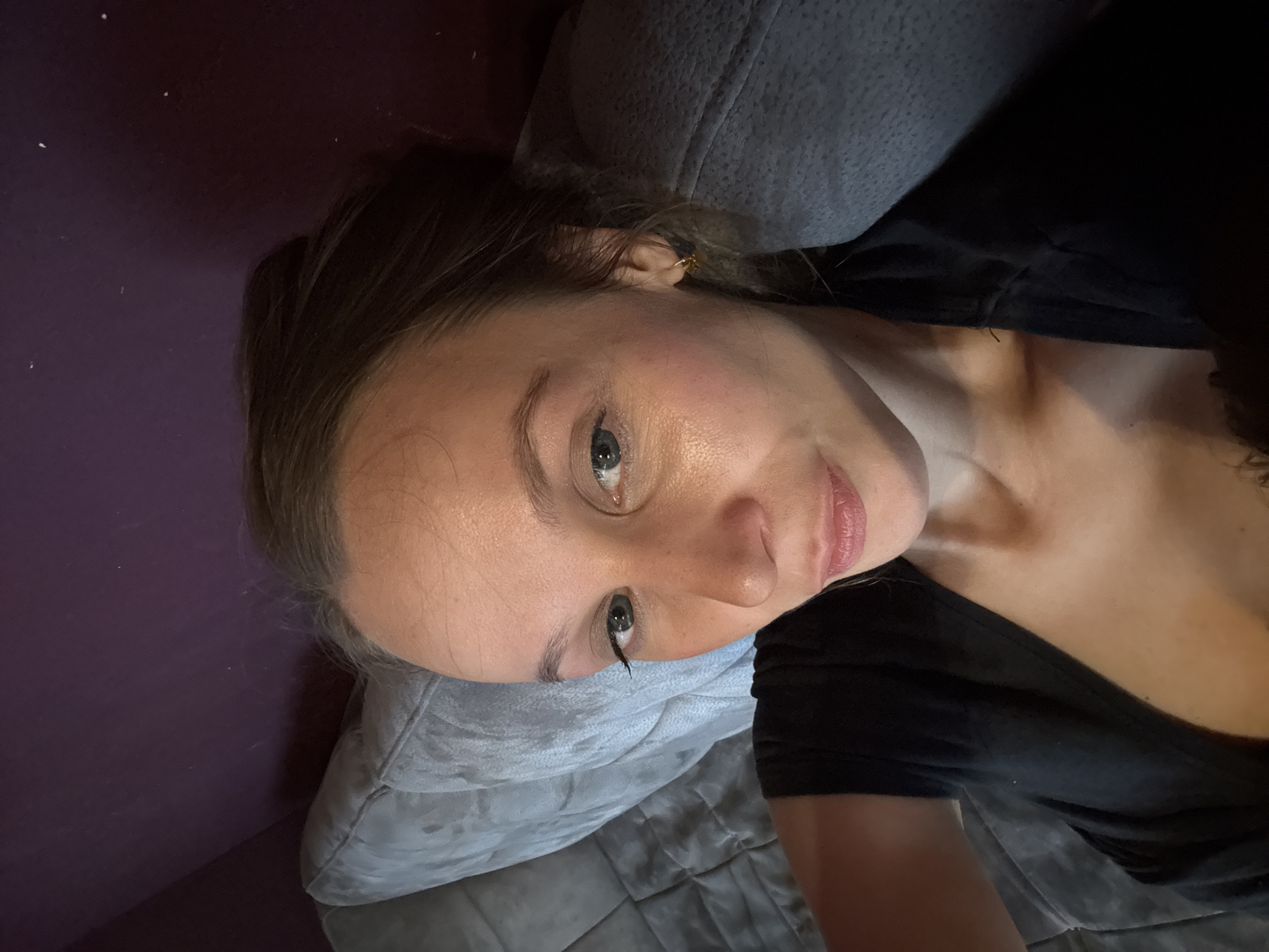 A woman is taking a selfie indoors. She is lying on a grey couch with a purple wall behind her. She is wearing a black shirt and has light brown hair pulled back. She is wearing a small gold earring in her right ear. Her makeup includes mascara and lip gloss. The image is taken from a high angle.