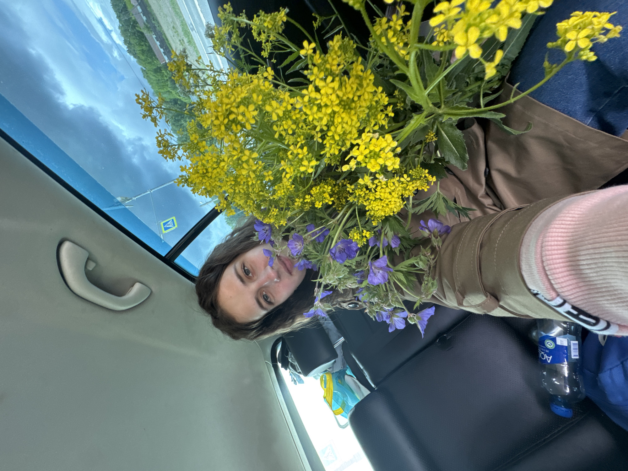A woman is taking a selfie in the back seat of a car. She is surrounded by a bouquet of yellow and purple wildflowers. The woman is wearing a brown jacket and a pink sock. There is a bottle of water and a blue bag next to her on the seat. Through the car window, trees and a road can be seen. A road sign is visible in the reflection of the window. Based on the sign, the location is in Zachernaya, Russia.