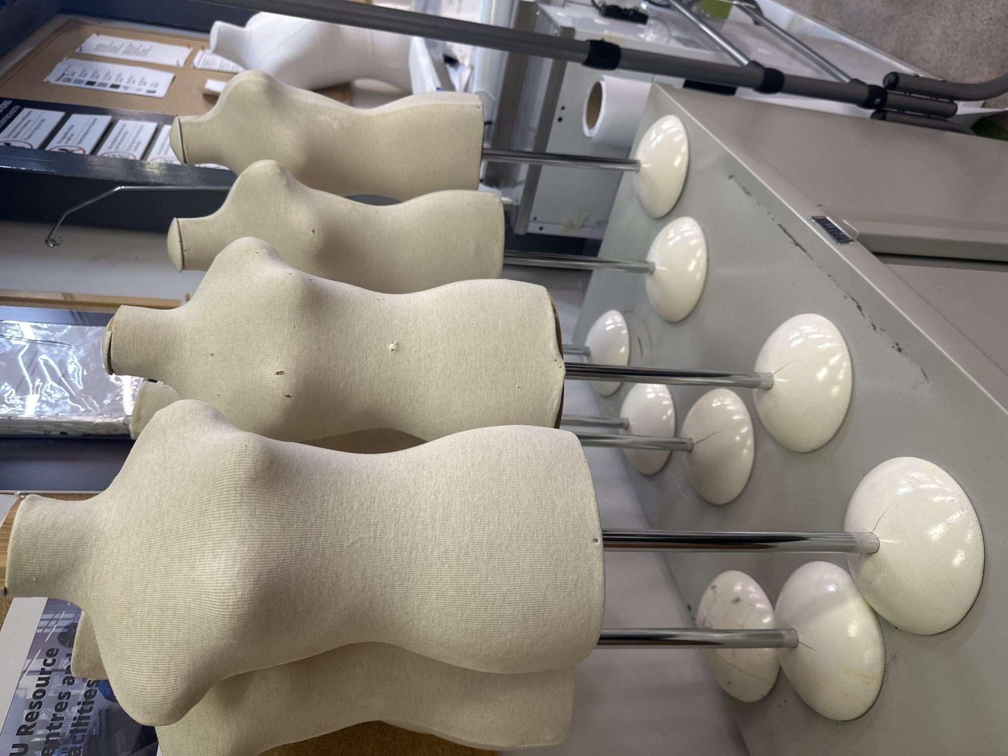 The image displays a row of five beige dress forms arranged on a metal surface. Each form is mounted on a chrome pole that connects to a white, circular base on the surface. A gray metal structure is visible in the background behind the dress forms. The city and specific location are unknown.
