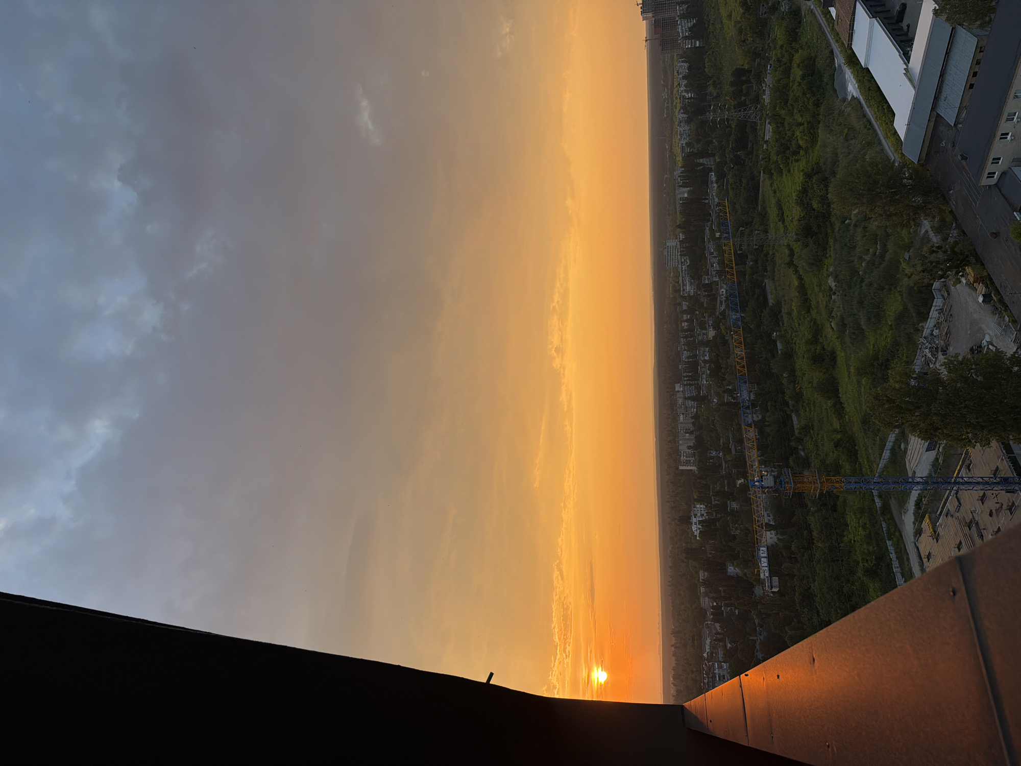 The image shows a sunset over a city, likely Kyiv, Ukraine, from a high vantage point. The sun is low on the horizon, casting a warm orange glow across the sky and the landscape. The foreground is dominated by a dark, angled structure, possibly a building facade or a balcony railing, which partially obscures the view.
Below the horizon, a sprawling cityscape unfolds, characterized by a mix of residential buildings, some appearing to be Soviet-era apartment blocks, and industrial structures. Greenery is abundant, with trees and vegetation interspersed throughout the urban environment. A prominent construction crane, painted in blue and yellow, stands out against the landscape, indicating ongoing development in the area. Electrical pylons are also visible, suggesting the presence of power infrastructure.
The sky is a gradient of warm oranges and yellows near the horizon, transitioning to cooler grays and blues as it ascends. Clouds are present, some appearing thin and wispy, while others are darker and more diffused. A few small birds are visible as tiny specks in the sky. The overall impression is a tranquil, late-afternoon or early-evening scene in an urban setting.