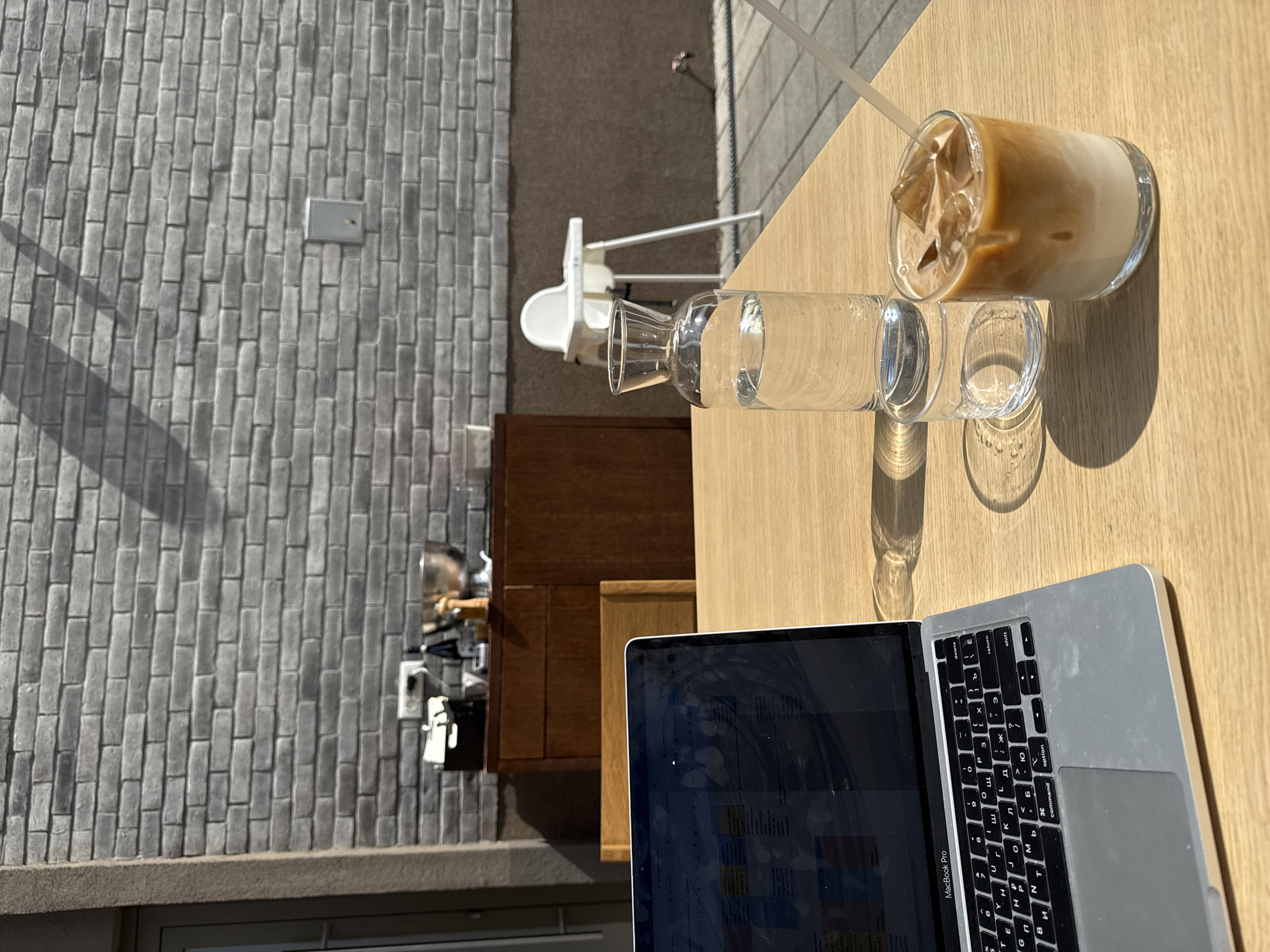This is an overhead shot of an outdoor cafe table, likely in Belgrade, Serbia, given the provided context and the Cyrillic keyboard layout. The image is bathed in bright sunlight, casting sharp shadows on the light wooden tabletop, suggesting it's either morning or afternoon on a clear day.
In the foreground, a silver MacBook Pro laptop is open, displaying what appears to be a spreadsheet or document on its screen. The keyboard prominently features both Latin and Cyrillic characters, with visible keys such as "delete," "return," "shift," "option," "command," and Cyrillic letters like "щ," "л," "д," "м," and others.
Next to the laptop, two glasses are placed. One is a tall glass filled with iced coffee, showing distinct layers of brown coffee over white milk, with ice cubes and a clear straw. Beside it is a clear glass carafe, filled with water, accompanied by an empty drinking glass.
The background reveals an outdoor setting. To the left, a textured grey brick wall extends vertically. Beyond the table, a dark, possibly carpeted, ground is visible, along with a white high chair, indicating a family-friendly cafe environment. Further back, wooden cabinets or furniture are partially seen, and a blurred reflection, possibly from a mirror or window, hints at an indoor area.
The scene depicts someone working or relaxing outdoors, enjoying beverages at a cafe. The strong sunlight and the presence of cold drinks contribute to a serene and productive atmosphere.