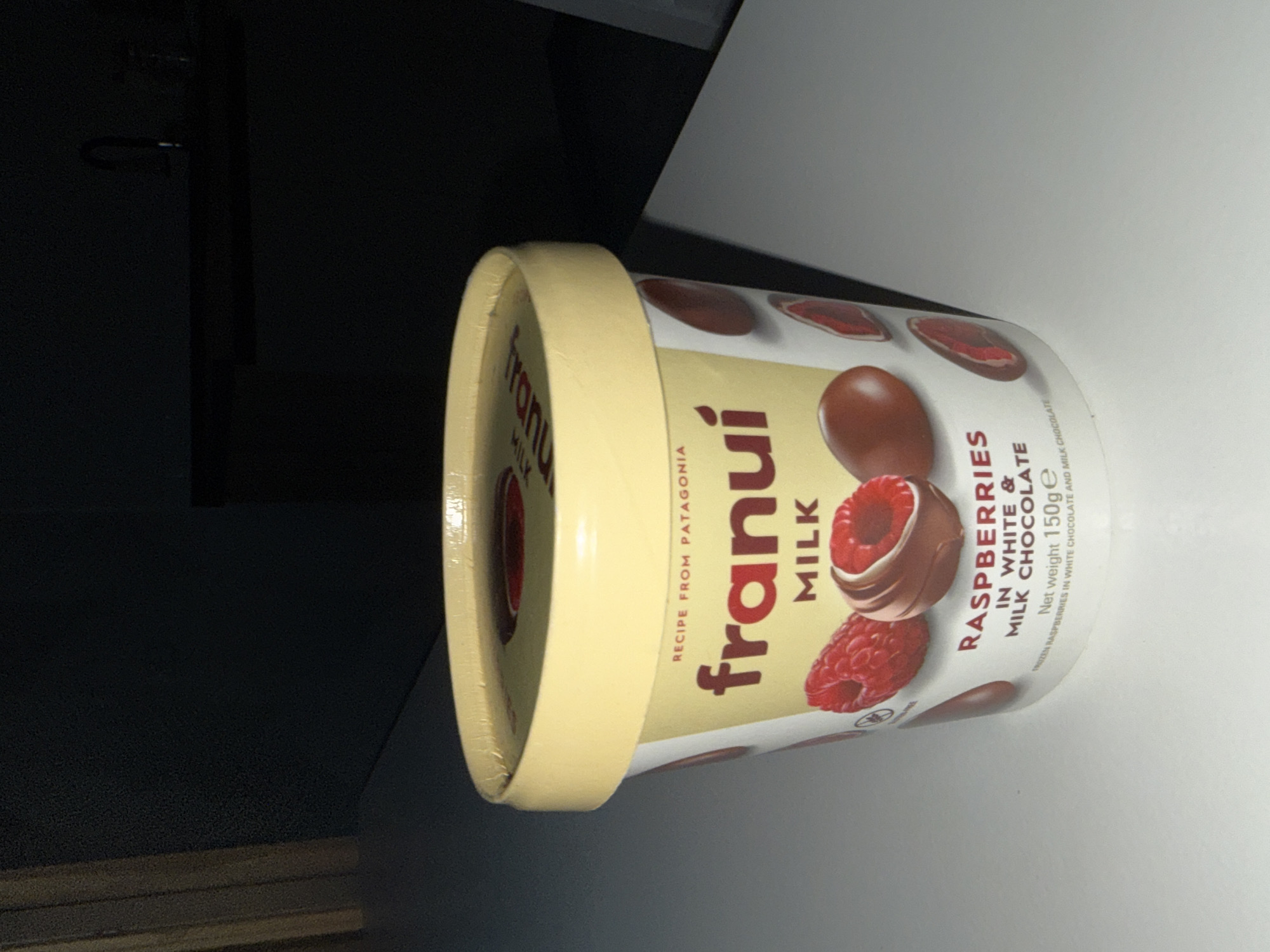The media file is a close-up, slightly tilted shot of a round, pint-sized container of "Frunui" brand milk chocolate with raspberries. The container is predominantly white with a yellow lid. The design features red raspberries and brown chocolate pieces, along with the brand name "Frunui" in a stylized font. Text on the container indicates it is a "RECIPE FROM PATAGONIA," contains "RASPBERRIES IN WHITE & MILK CHOCOLATE," and has a "Net weight 150g." The image has a somewhat dramatic, low-light setting, with shadows obscuring much of the background, suggesting an indoor environment, possibly a countertop. The focus is sharp on the product itself, highlighting the packaging details. There are no people or animals visible in the image.