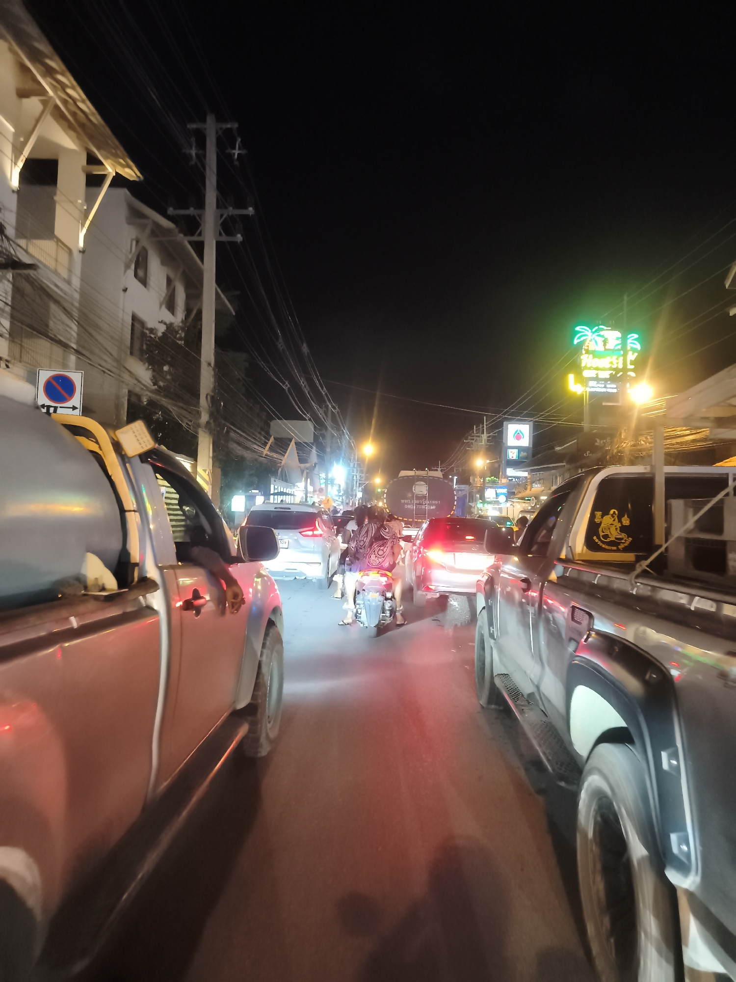 The image depicts a nighttime scene of a congested street in the Ko Samui District, Thailand, likely a busy thoroughfare with commercial establishments. Several vehicles, including pickup trucks, cars, and a motorcycle, are stopped in traffic. The vehicles appear to be modern models. The lighting suggests it's after dark, with artificial lights from buildings and street lamps illuminating the scene. Some buildings are visible on the left, featuring architectural elements and outdoor lighting. A prominent neon sign with palm tree imagery is visible in the background, indicating a commercial area. The road surface is asphalt. The presence of multiple vehicles and the surrounding infrastructure suggest an urban or semi-urban environment.