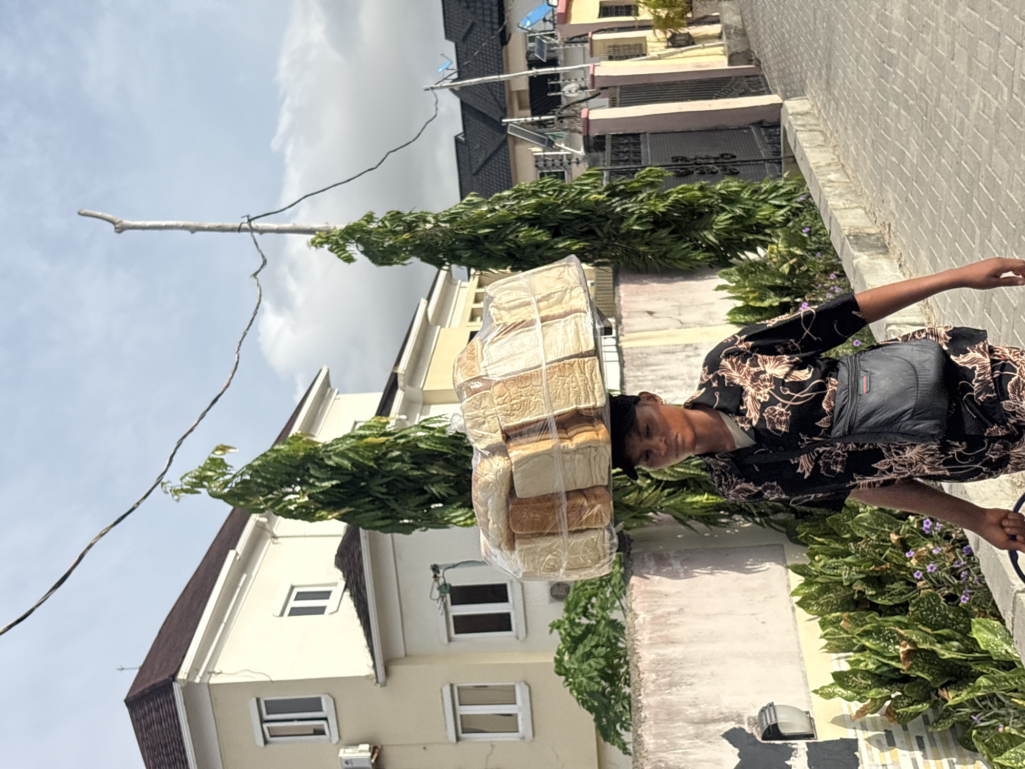 The image shows a man walking in Igboefon, Nigeria with several loaves of bread stacked on his head. He is wearing a black floral shirt and carrying a black bag on his shoulder. The loaves of bread are wrapped in clear plastic. The background includes buildings, trees, and a sky with clouds. A powerline and a wooden pole are also visible.