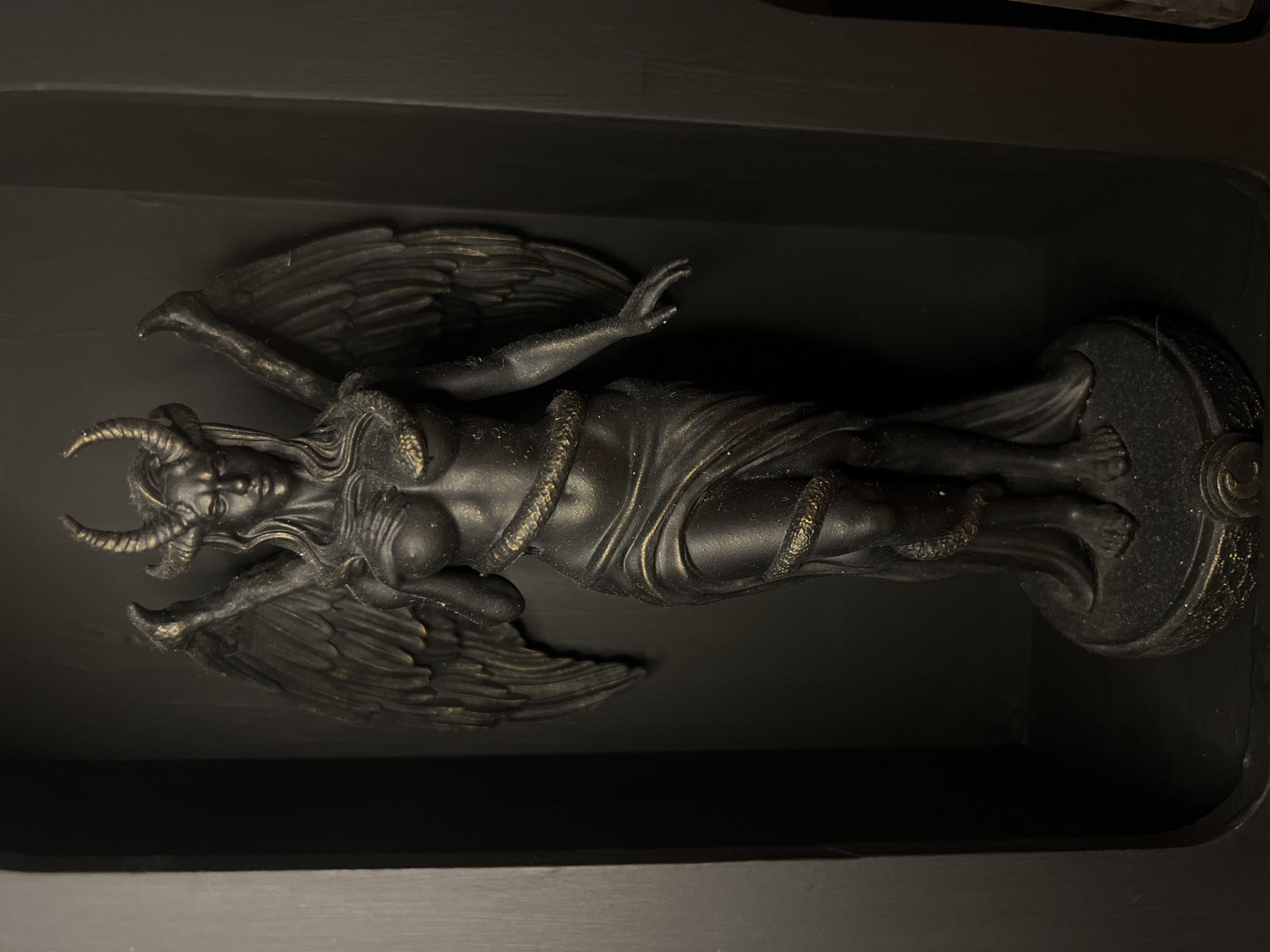 A dark, detailed statue of a winged female figure with horns and a serpent wrapped around her is displayed within a dark, recessed shelf. The statue is predominantly black with subtle gold accents on the horns, wings, and parts of the serpent and drapery. The figure has long, flowing hair, and its expression is stern. Its wings are spread, and its arms are positioned slightly outwards, with one hand gently extended. The serpent is coiled from the base of the statue up around the figure's torso and neck. The base of the statue is also dark and textured, with visible gold detailing at the front. The shelf is dark, appearing to be made of wood, and the lighting is dim, casting shadows that enhance the statue's dramatic appearance. The overall impression is one of dark fantasy or gothic art.