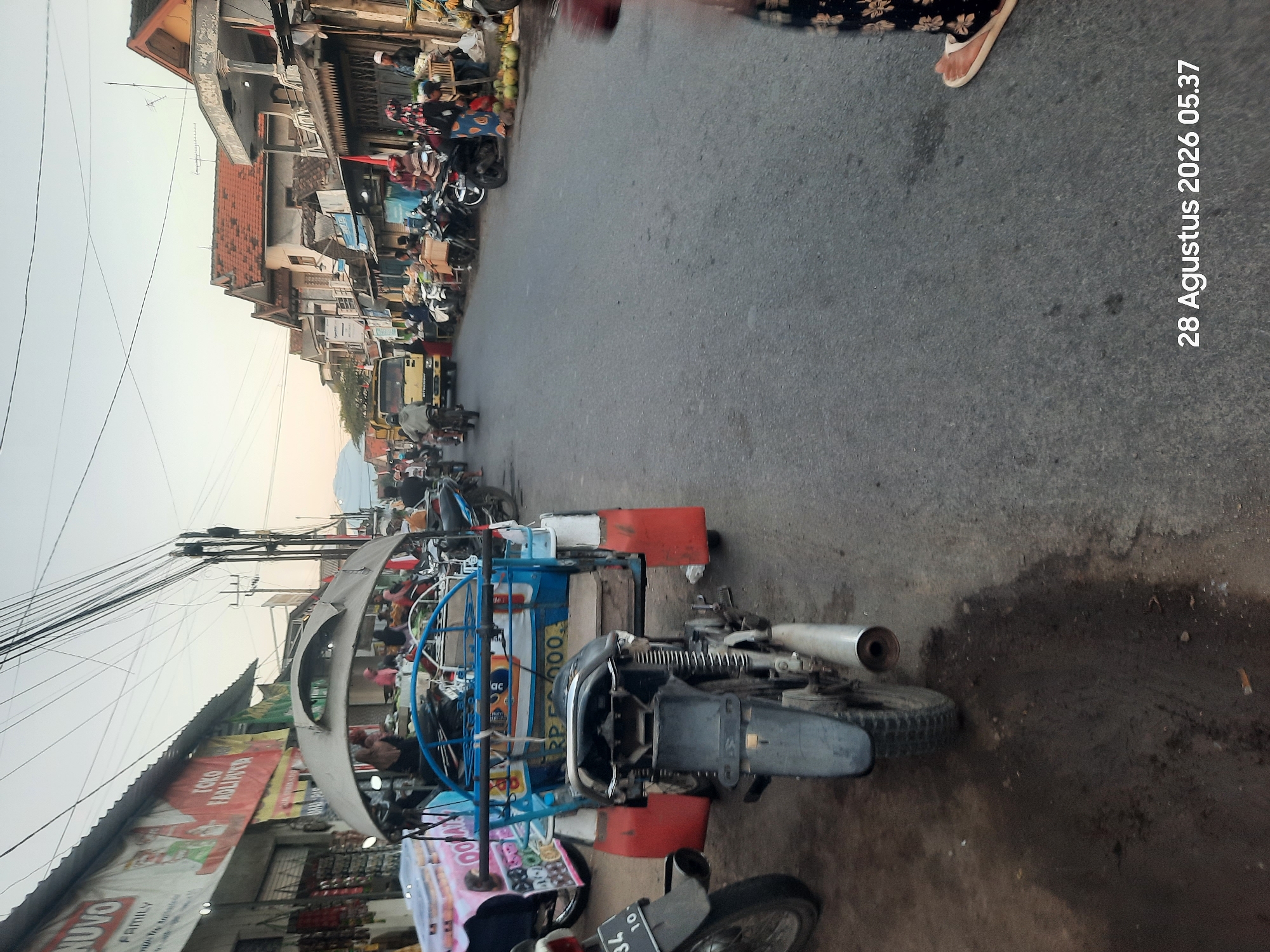 The image captures a street scene in Pajarakan Kulon, Indonesia, likely during a market day or a busy period. The foreground is dominated by the rear view of a blue and red motorized tricycle, with its black seat and metallic exhaust pipe clearly visible. To the left, the rear wheel and fender of a motorcycle are partially visible.
The street is lined with buildings that appear to be shops or market stalls. Some of these structures have corrugated metal roofs, and banners with text are displayed on their facades, suggesting commercial activity. In the middle ground, several motorcycles are parked along the side of the road, and people are engaged in various activities. Some individuals are seen standing, while others are on motorcycles. A yellow truck is also present, moving along the street.
Further down the street, more shops and buildings are visible, with people milling about. The overall atmosphere suggests a lively and active urban environment. The sky in the background is clear, indicating fair weather. A timestamp in the bottom right corner reads "28 Agustus 2026 05.37", indicating the date and time the photograph was taken.