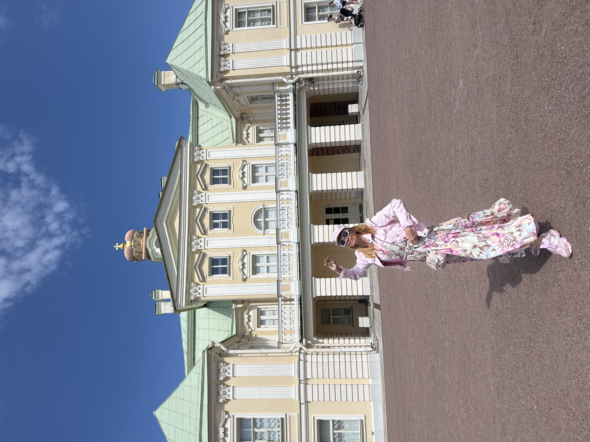 A woman stands in front of a grand, yellow building with a light green roof, possibly a palace or mansion, under a clear blue sky with some white clouds. The building features ornate white trim, columns, and a balcony. A prominent crown adorns the central dome of the building. The woman is wearing a pink denim jacket over a floral-print dress, a pink baseball cap, sunglasses, and pink sneakers. She is holding her right hand up, possibly waving. A small, decorative purse is slung across her shoulder. The ground in front of the building is a reddish-brown gravel. In the background, to the right, a stroller with two people can be seen. The scene appears to be the exterior of the Catherine Palace (also known as the Grand Palace of Tsarskoye Selo), a famous landmark located in the city of Lomonosov, Russia.