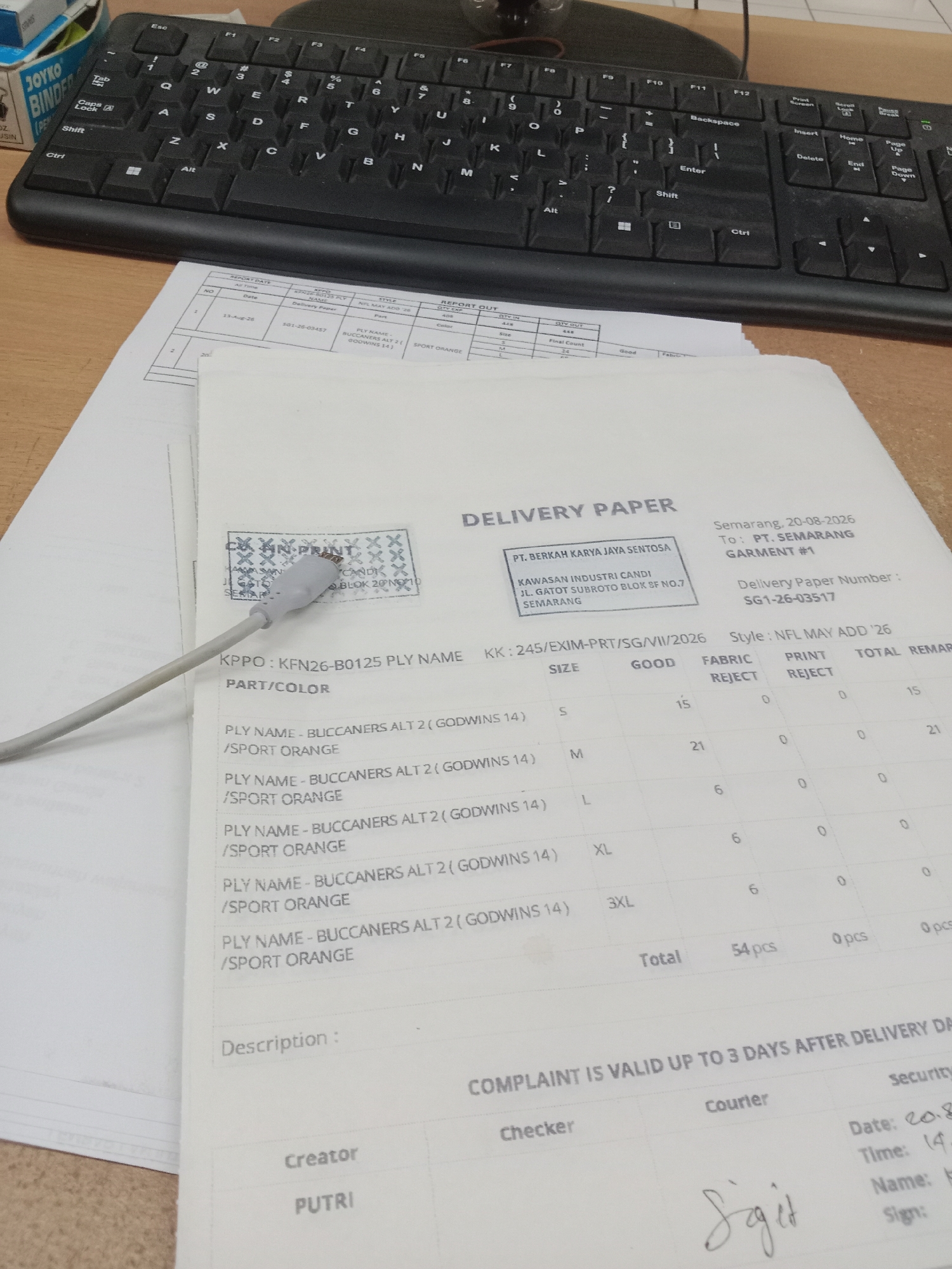 The image shows a close-up of a desk with a keyboard and a delivery paper. The delivery paper is from "PT. BERKAH KARYA JAYA SENTOSA" located in Semarang, Indonesia. The paper details a delivery of "KPPO: KFN26-B0125 PLY NAME" with various sizes and quantities, and notes a delivery number of "SG1-26-03517". The date of the delivery paper is August 20, 2026. A grey USB cable is resting on the paper, partially obscuring a stamp that reads "PRINT". The environment appears to be an office or workspace.