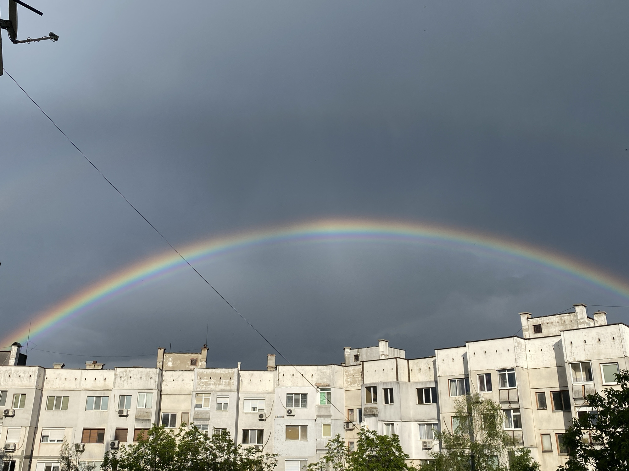 The image shows a rainbow arched over a drab, concrete residential building in Pleven, Bulgaria. The sky is overcast with dark gray clouds, indicating a recent rain. In the foreground, treetops are visible, partially obscuring the lower part of the building. The building is a multi-story structure with several rows of windows and air conditioning units attached to the facade. A utility pole with wires extending across the top left corner of the frame. The rainbow is vibrant, contrasting sharply with the somber sky and the utilitarian architecture. The overall scene presents a juxtaposition of natural beauty against an urban backdrop.