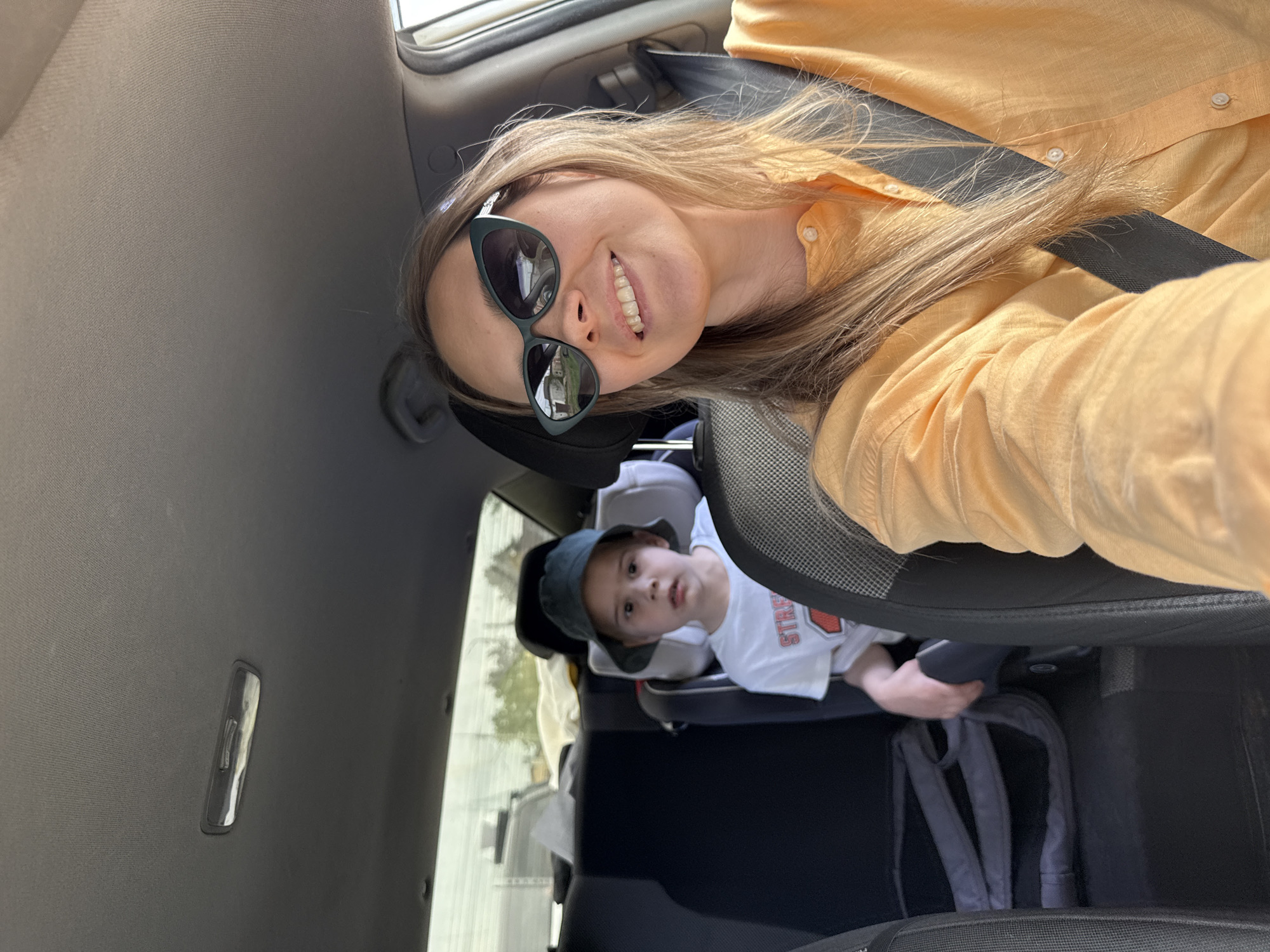 The image shows a woman with sunglasses and a yellow shirt sitting in the driver's seat of a car, wearing a seatbelt. In the back seat, a young child is secured in a car seat, wearing a white shirt and a dark green hat. A grey backpack rests behind the child's seat. The interior of the car is visible, including the headrest of the driver's seat and the car's ceiling. The location is unknown, but the prompt suggests it may be Chaikovsky, Russia.