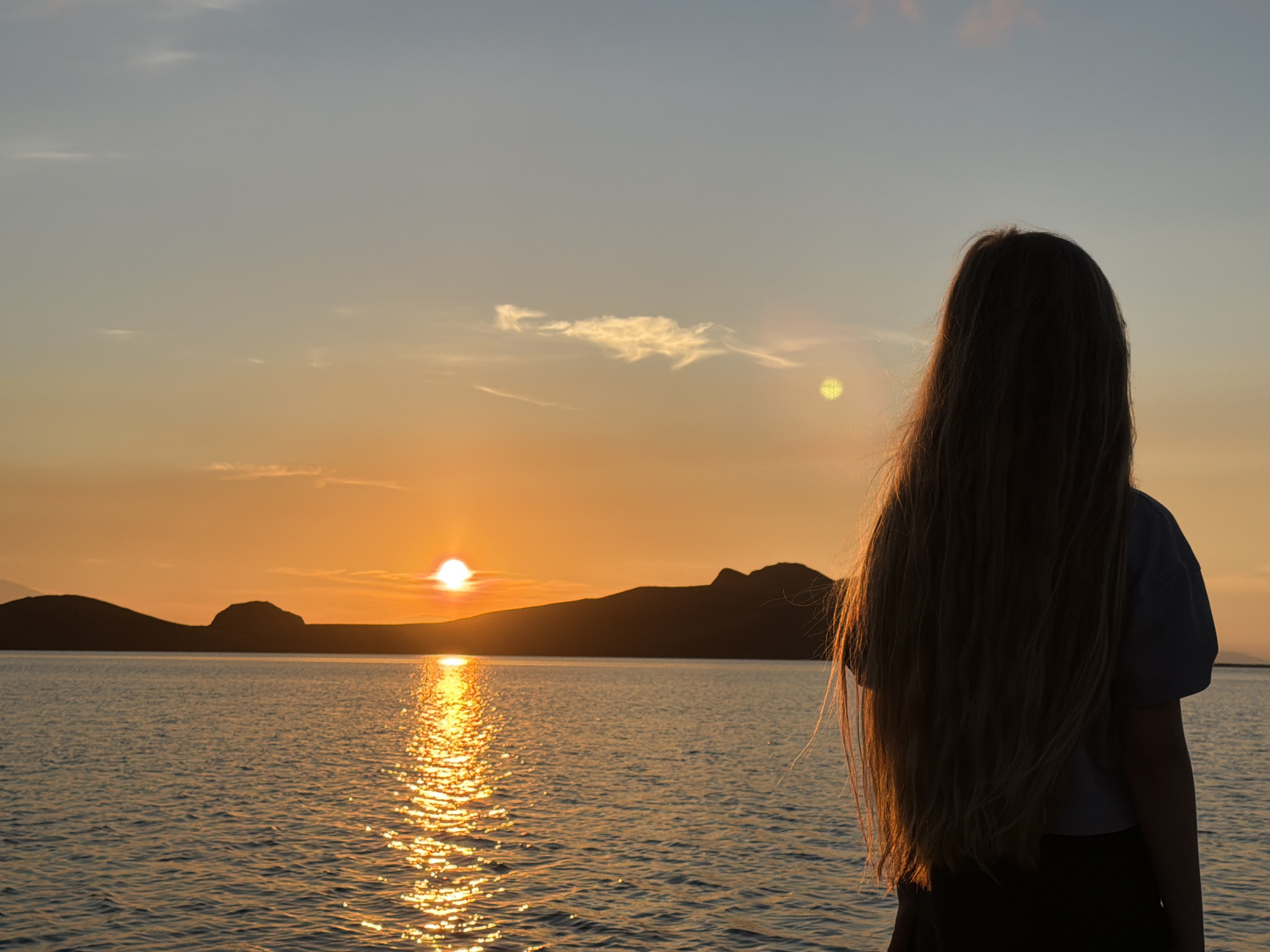 A person with long, blonde hair stands facing a body of water during sunset. The sun is low on the horizon, casting a golden glow across the water and silhouetting distant hills. The sky is a gradient of orange and blue, with a few wispy clouds. The person is wearing a dark shirt and appears to be observing the sunset. The scene is peaceful and serene, evoking a sense of calm and reflection. The location is unknown.