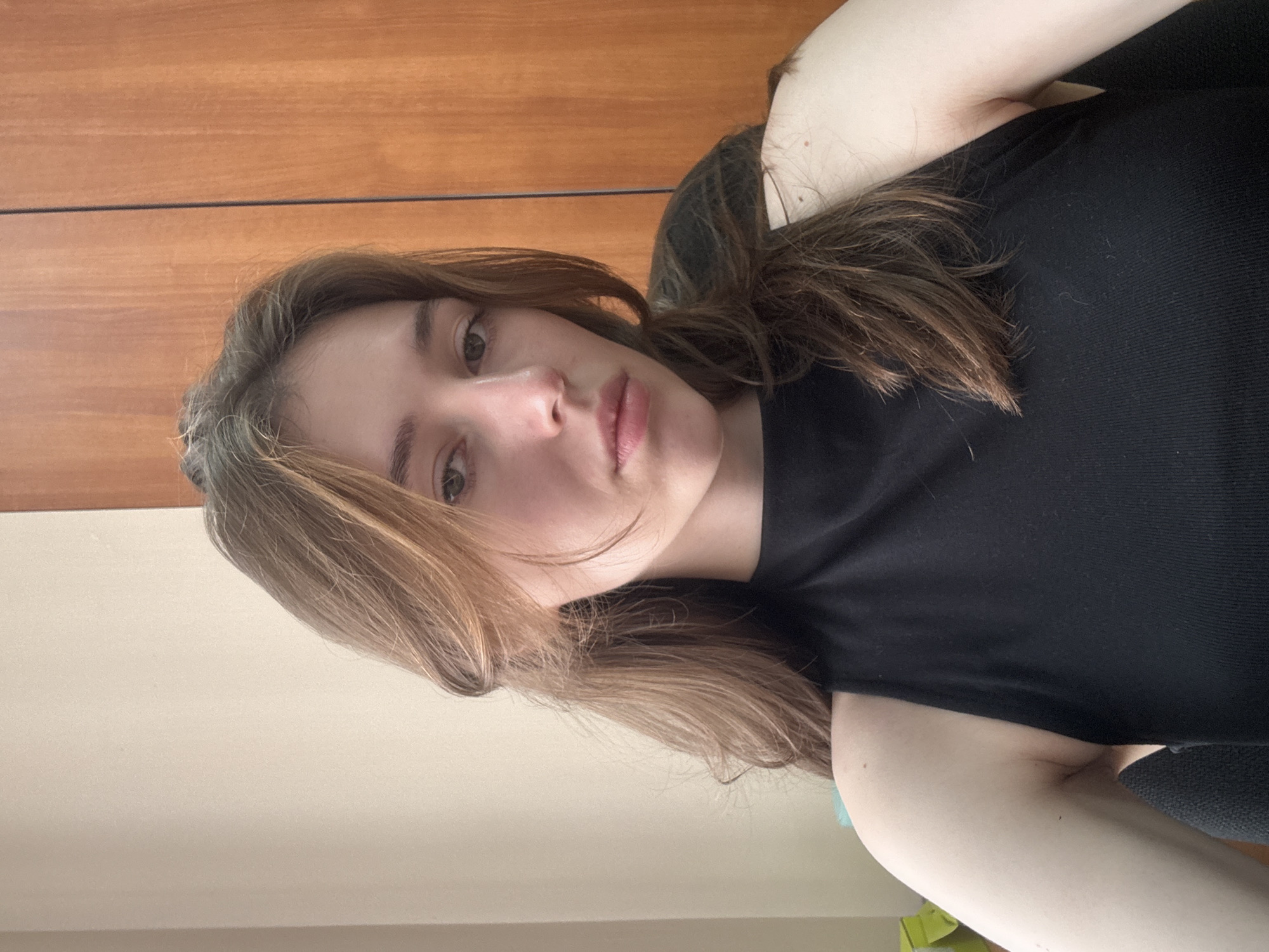 A young woman with long, brown hair is looking at the camera. She is wearing a black sleeveless shirt. The background consists of a beige wall and a wooden cabinet with vertical lines. The scene appears to be an indoor setting, likely an office or a home. There are no discernible landmarks or indications that would pinpoint the location to the city of Perm, Russia.
