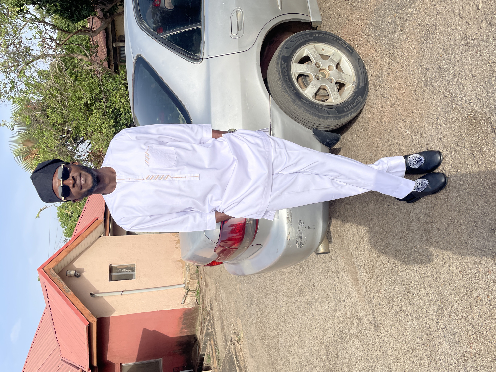 A man in a white outfit and sunglasses stands next to a silver car on a paved surface. He wears a black cap and black shoes with decorative stitching. The car is parked near a building with a red roof and beige walls. Trees and vegetation are visible in the background. The setting is outdoors, on a sunny day.