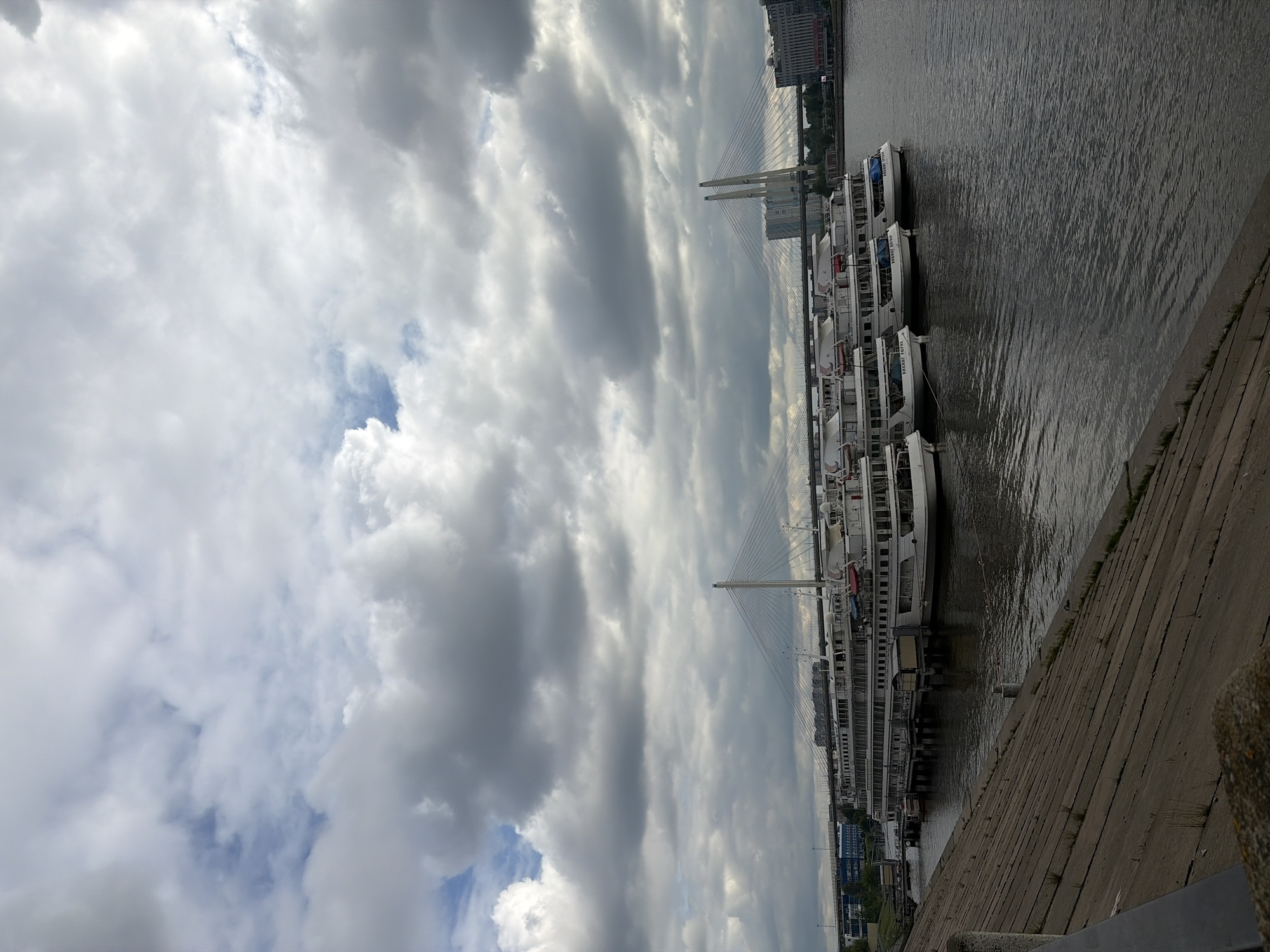 The image depicts a cloudy sky over the Neva River in Saint Petersburg, Russia. A cable-stayed bridge, likely the Volodarsky Bridge or a similar structure, is visible in the distance. A row of docked white passenger boats lines the riverbank. A concrete embankment stretches along the foreground. No people are visible.