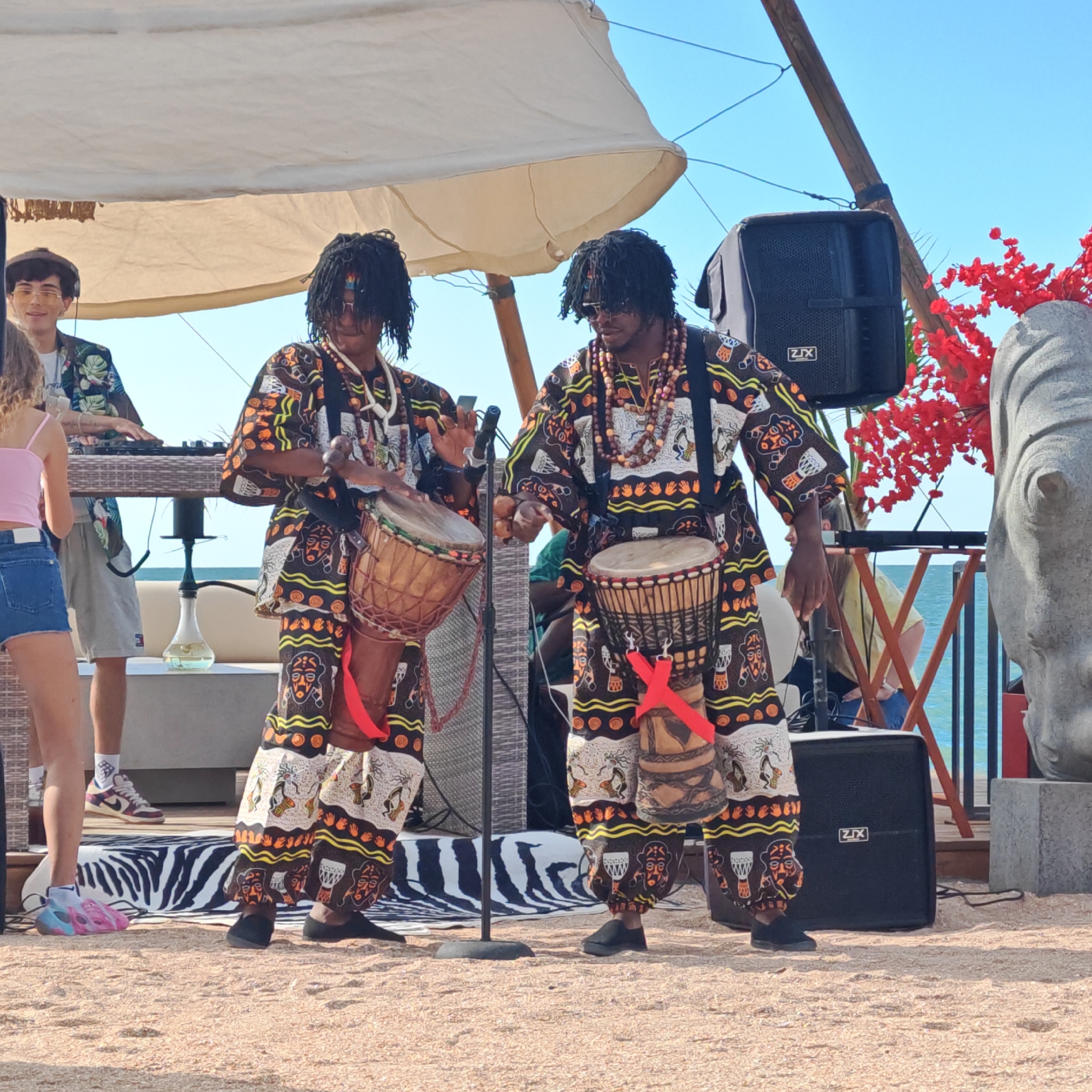 The image depicts an outdoor performance on a sandy beach, likely near the sea given the distant blue water and clear sky. Two individuals are prominently featured, dressed in vibrant, patterned clothing reminiscent of traditional African attire. Both are wearing elaborate beaded necklaces and have dark, braided hair. They are actively playing hand drums, or djembe drums, which are strapped to their bodies. Their stance and the way they are holding their drumsticks suggest they are in the midst of a rhythmic performance.
To their left, a person wearing a floral shirt is operating a DJ console, with a set of turntables visible on a table. A woman in a pink tank top and denim shorts stands near the DJ, observing the scene. In the background, a large speaker is visible, and to the right, a grey statue that appears to be a rhinoceros head is partially visible, along with a decorative red tree. The scene is set under a large, off-white canopy, providing shade. The overall atmosphere appears festive and energetic, suggesting a daytime event given the bright sunlight. The sandy ground and the presence of the sea indicate a beach or coastal location. A hookah pipe is also visible on a table behind the DJ. The speaker has "ZLX" visible on it.