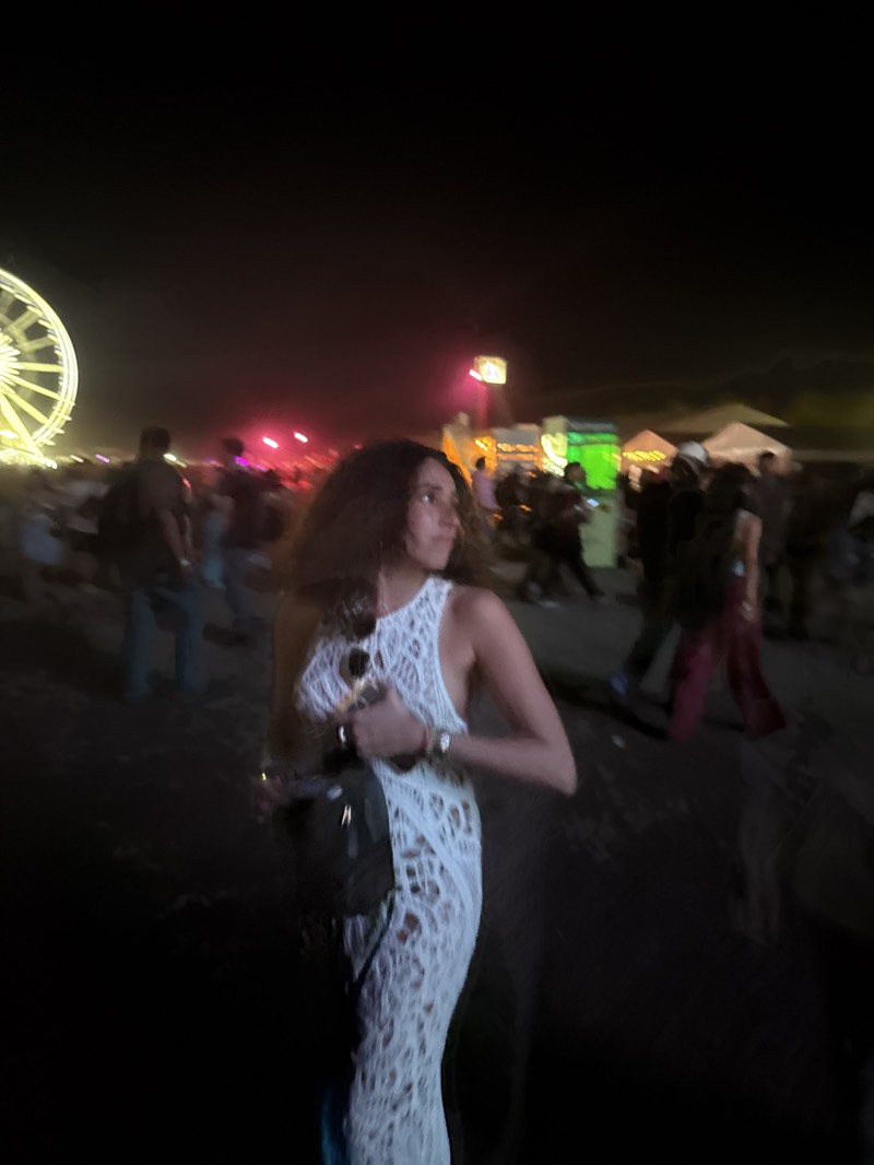The image shows a woman wearing a white, sleeveless, floor-length dress. She has long, dark hair and is wearing a watch on her left wrist. She is carrying a dark-colored bag on her shoulder and sunglasses are placed on her top. The background suggests a crowded outdoor event at night. A Ferris wheel is visible on the left side of the image. Other people are present and there are food stalls and lights. Based on the background, it appears to be the Coachella Valley Music and Arts Festival in Indio, United States.