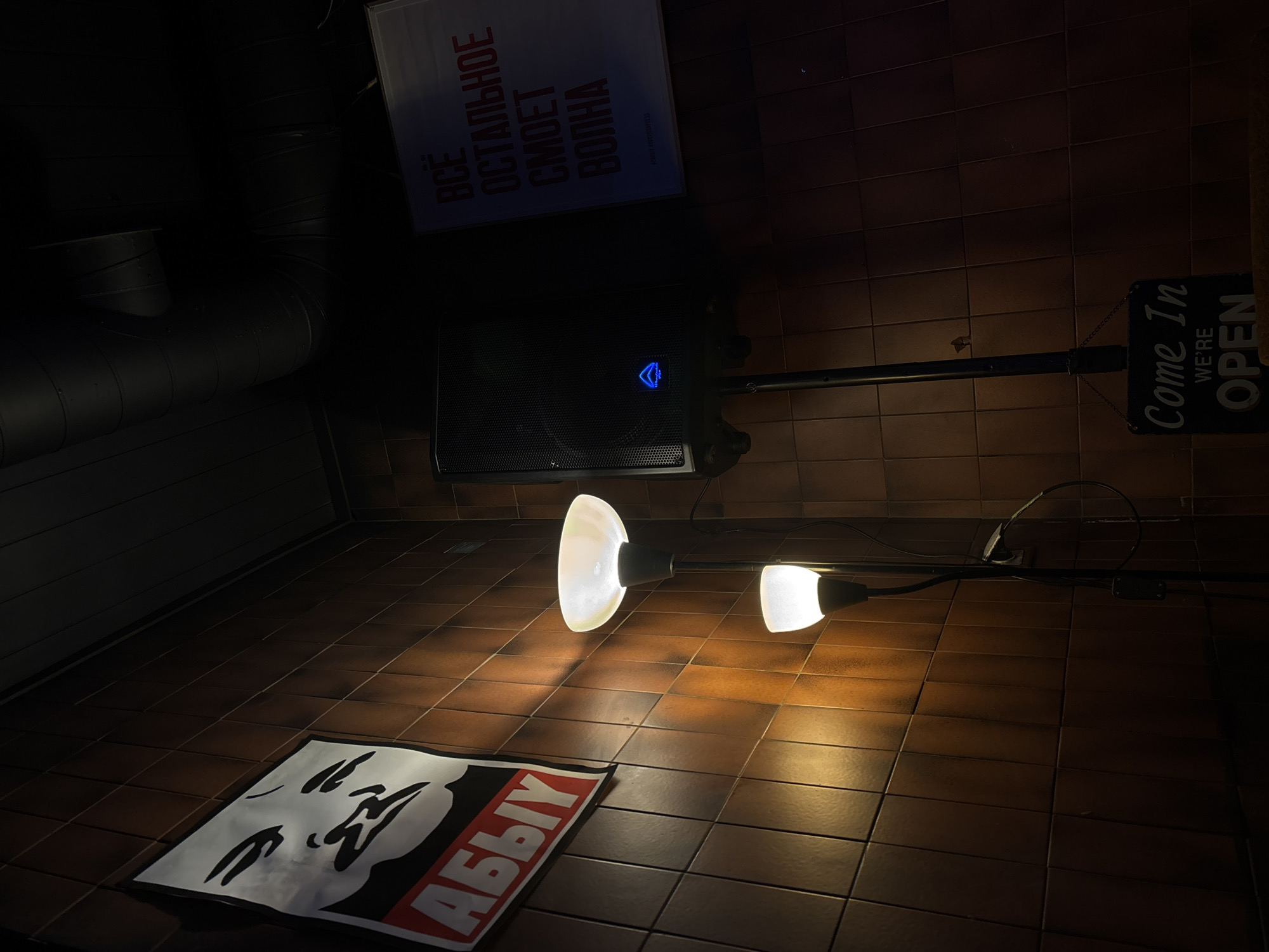 The image shows a scene indoors, likely a basement or a dimly lit interior space. The floor is covered in brown tiles. A black and white poster with a face and Cyrillic text ("АБЫУ") lies on the floor. Above it, two white, lamp-like lights illuminate the space, attached to a horizontal bar. Further up, a black speaker with a blue logo is mounted on the wall. Above the speaker, there is a poster with Cyrillic text in red. To the right of the speaker, a "Come In We're Open" sign hangs. A dark pipe runs along the upper left corner of the image. The overall lighting is dim, with the main light sources being the two lamp lights. No people are visible. The location is suggested to be in Belgrade, Serbia, due to the presence of Cyrillic text.