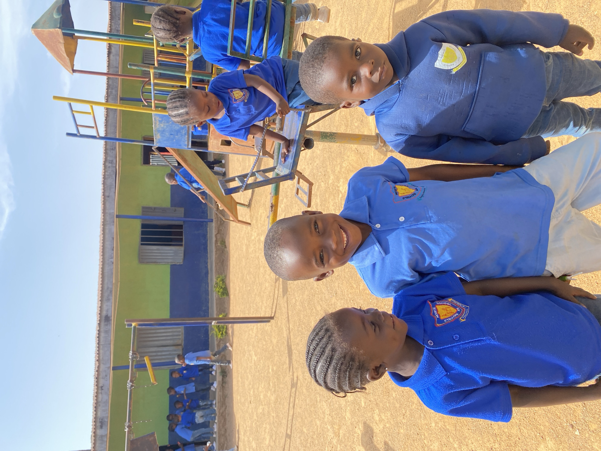 The image shows a group of children in blue uniforms on a playground in Gura Top, Nigeria. They are likely students. The playground has a metal structure with bars, ladders, and a swing-like seesaw. In the background is a two-story building with green and blue painted walls. The ground is a light-colored sandy soil. The sky is pale blue. The scene appears to be outdoors in daylight.