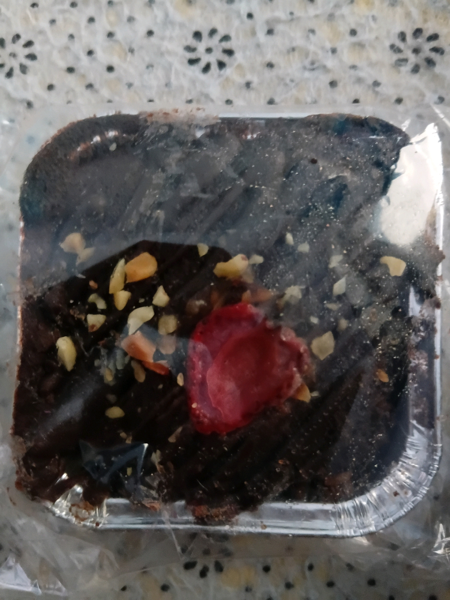 A single, square-shaped brownie is presented in a shiny, fluted aluminum foil container. The brownie is dark brown and appears dense, with a moist, fudgy texture visible through a clear plastic wrap. Scattered across the top of the brownie are chopped nuts, which are light yellow or tan in color. A single, bright red, petal-like topping, possibly a dried fruit or candy, is positioned slightly off-center on the brownie. The background consists of a speckled, textured surface, likely a tablecloth or placemat, with a floral pattern of dark, circular shapes. The overall lighting is neutral. The image provides no information about the location in Tanggul Wetan, Indonesia, or any people or actions.