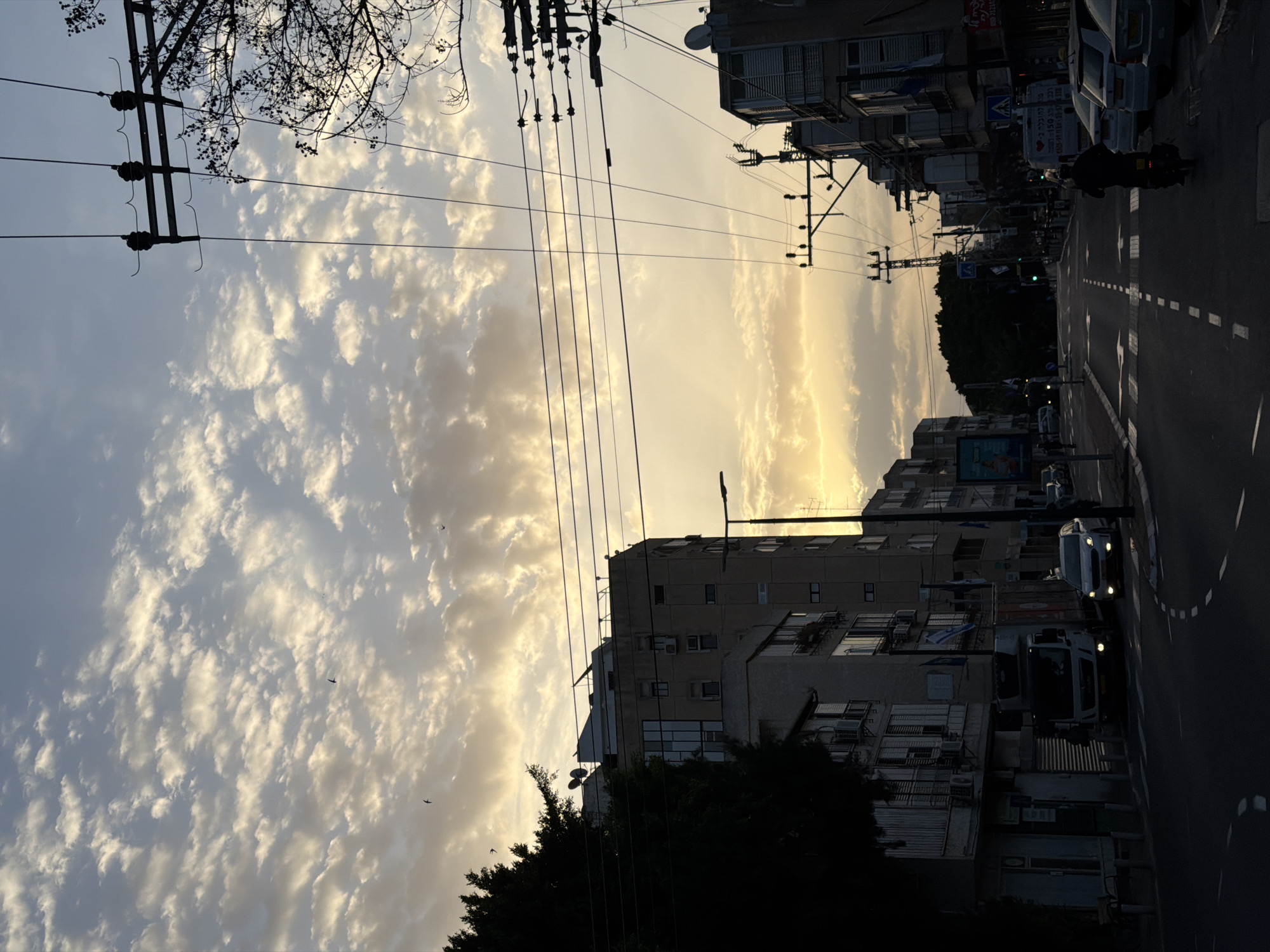 The image shows a street scene in Ramat Gan, Israel, at sunset. The sky is filled with scattered clouds, illuminated by the setting sun. Overhead electrical lines run across the sky. Buildings line both sides of the street. A street curves into the distance with traffic in the form of a motorbike and two cars. The buildings are multi-story, typical of urban residential areas. Some trees are visible in the foreground.