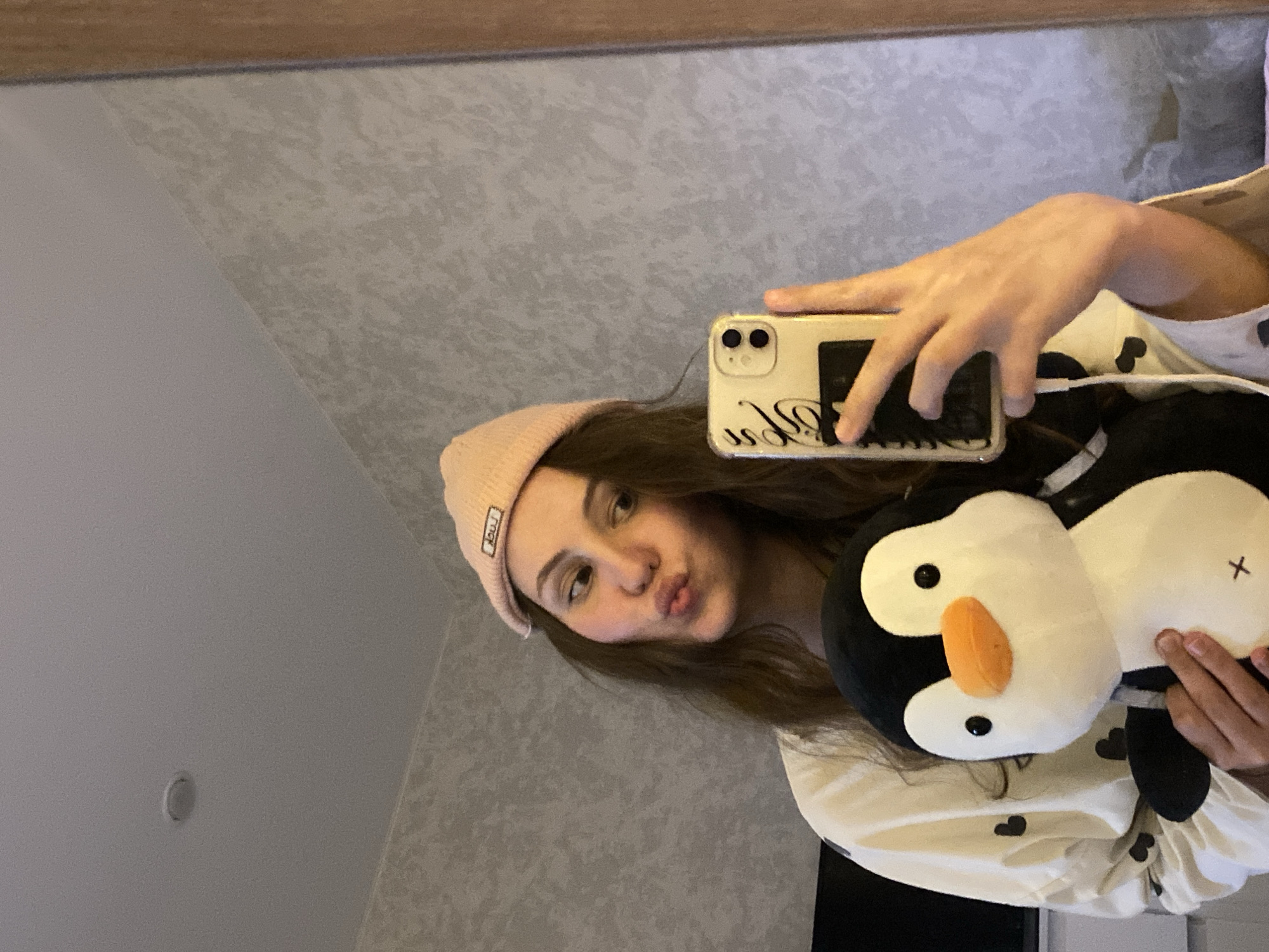 A young woman is taking a selfie in a mirror, holding a penguin stuffed animal. She is wearing a pink beanie and a white shirt with black hearts. The woman has brown hair and is making a kissing face. The phone she is using has a white case with black writing. The background is a light gray wall and ceiling. The location is Unknown, Unknown.
