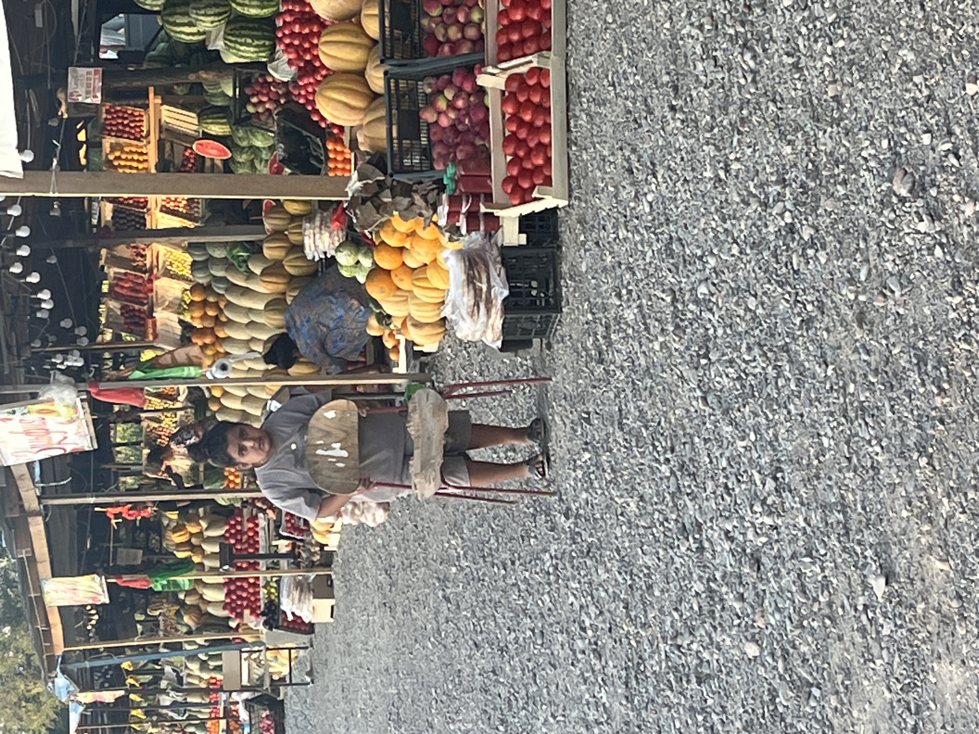 A young person stands in a fruit and vegetable market in an outdoor setting. The person is wearing a gray t-shirt and shorts and is holding a wooden object. They are standing on a gravel surface. In the background, stalls are filled with various produce, including watermelons, pumpkins, tomatoes, apples, and other fruits and vegetables. The market appears to be under a wooden canopy with lights strung overhead. Another person is visible in the background, partially obscured by the stalls. The overall impression is of a bustling market scene with a focus on fresh produce.