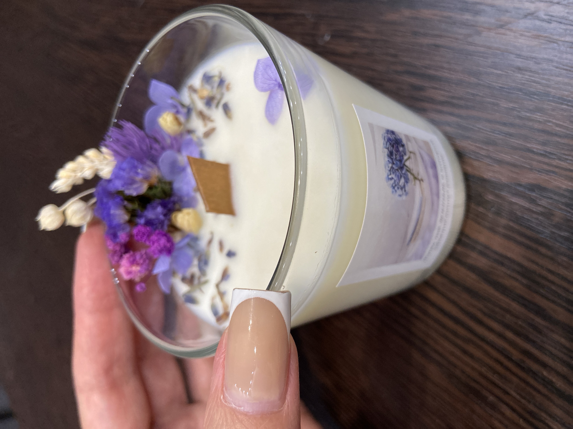 A close-up shot shows a hand with a French manicure holding a candle. The candle is in a clear glass jar and is topped with dried flowers and a wooden wick. The wax is a creamy white color and appears to have small dried flowers embedded in it. A label with a floral design is visible on the side of the jar. The background is a dark wooden surface. The image does not contain information that would allow for the determination of its location in Raevka, Russia.