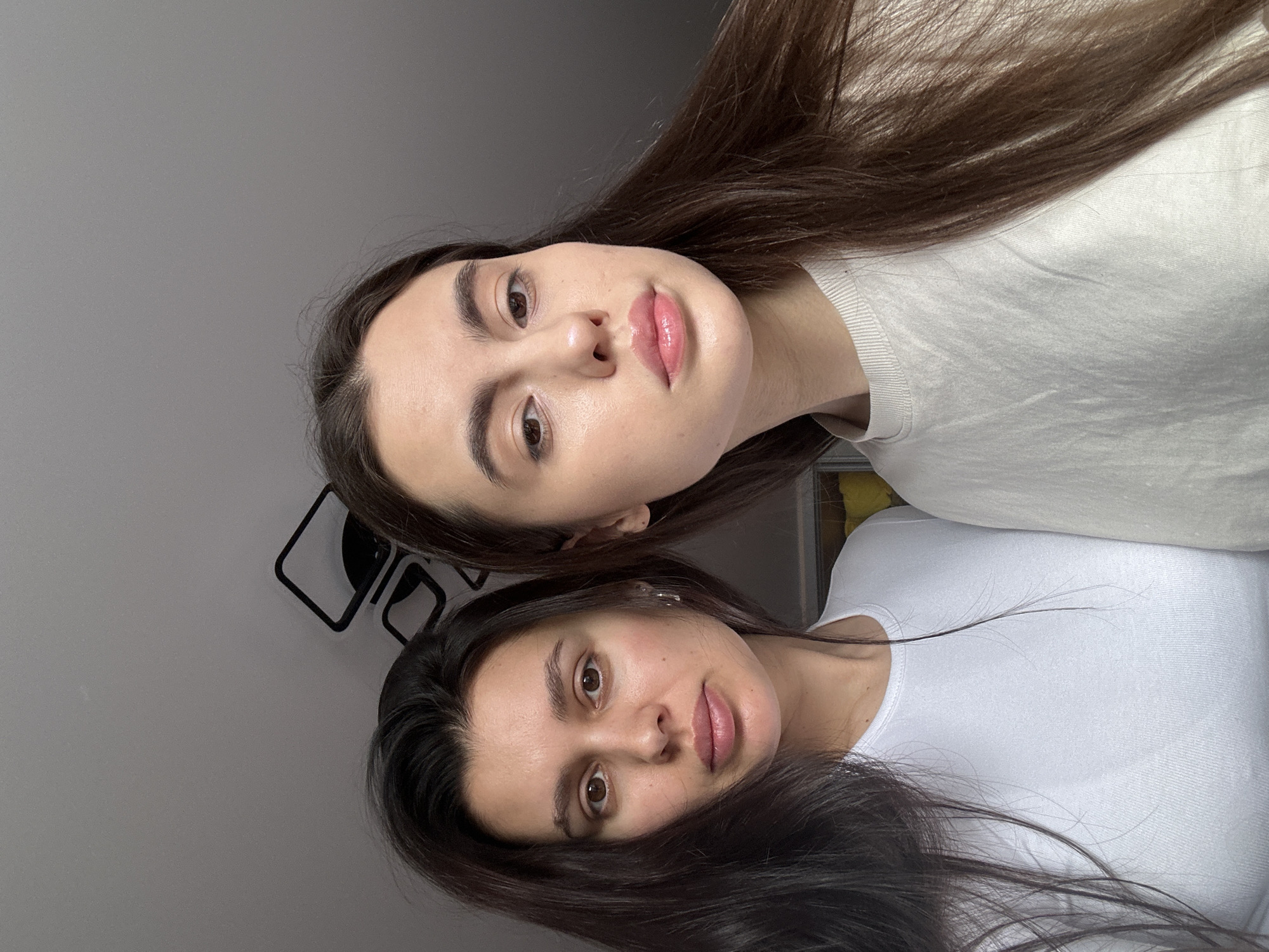 This image, captured in Chaikovsky, Russia, is a portrait featuring two young women posing for the camera. The photo is taken indoors against a plain, light gray wall.
The woman in the foreground, positioned slightly lower in the frame (if viewed correctly oriented), has long, dark brown hair that cascades over her shoulders. She wears a simple white crew-neck top. Her gaze is directed towards the camera, and she has a neutral expression with slightly parted lips. Her complexion is fair, and her eyebrows are dark and well-defined.
Behind and slightly above her is another woman, also with long, dark brown hair. She is wearing a light-colored (possibly off-white or light gray) ribbed long-sleeved top. Like the first woman, she looks directly at the camera with a neutral to slightly serene expression. Her lips are full and appear glossy.
In the upper left background, behind the women, a minimalist black geometric wall decoration is visible, consisting of square or rectangular outlines. The lighting is soft and even, suggesting either natural light from a window or well-diffused indoor lighting, illuminating their faces clearly. There is no visible text in the image. The scene conveys a calm and casual atmosphere.