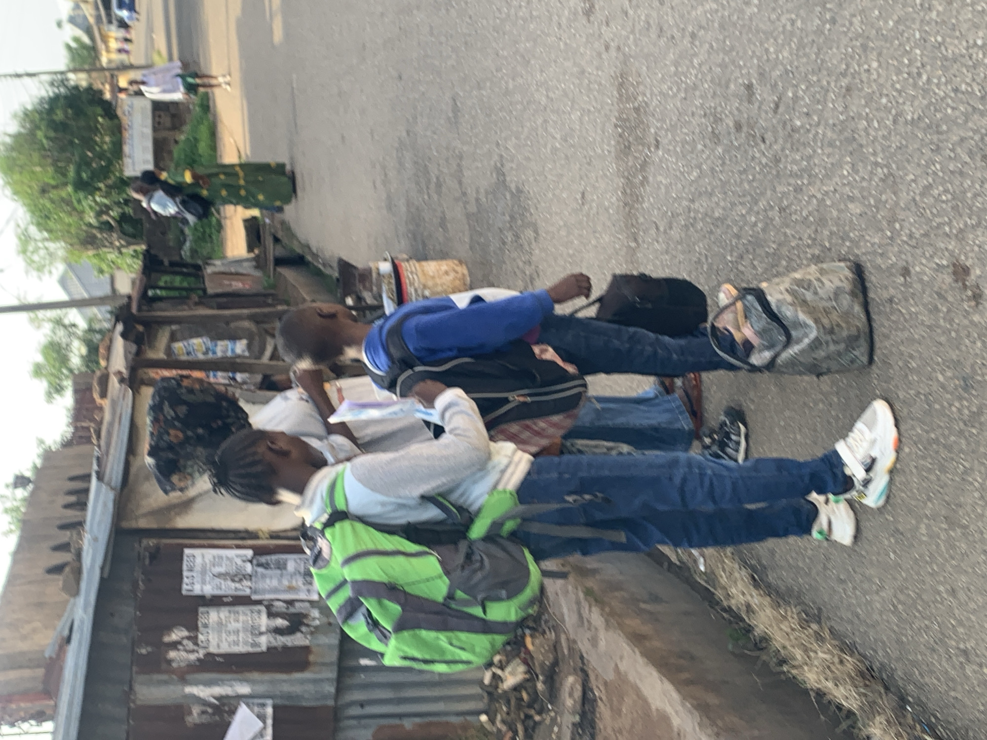 The image depicts a street scene in Jos, Nigeria, with several individuals and objects. In the foreground, two young people are seen standing. One, wearing a blue hooded sweatshirt and jeans, has a backpack. The other, in a light blue sweatshirt and jeans, carries a bright green backpack. Their attire suggests casual wear. They appear to be waiting or traveling, with several bags placed on the ground next to them, including a large, patterned duffel bag and a dark-colored suitcase.
Behind them, a small, weathered stall or kiosk is visible, constructed with corrugated metal and wood. This structure appears to be part of a marketplace or roadside vending area, with items displayed or stored within. A woman in a green patterned dress is seen near this stall, possibly interacting with someone or engaged in an activity related to the stall. Further in the background, trees and other structures are blurred, indicating the surrounding environment is urban or semi-urban.
The lighting suggests it is daytime, likely morning or afternoon, with sunlight casting shadows. The road surface is asphalt, and there are signs of vegetation and debris on the side of the road. The overall atmosphere seems to be that of a typical day in a bustling area. Some text is visible on posters or flyers attached to the metal siding of the stall, but it is too blurry to be legible. The focus of the image is on the young people and their belongings, possibly indicating travel or transit.