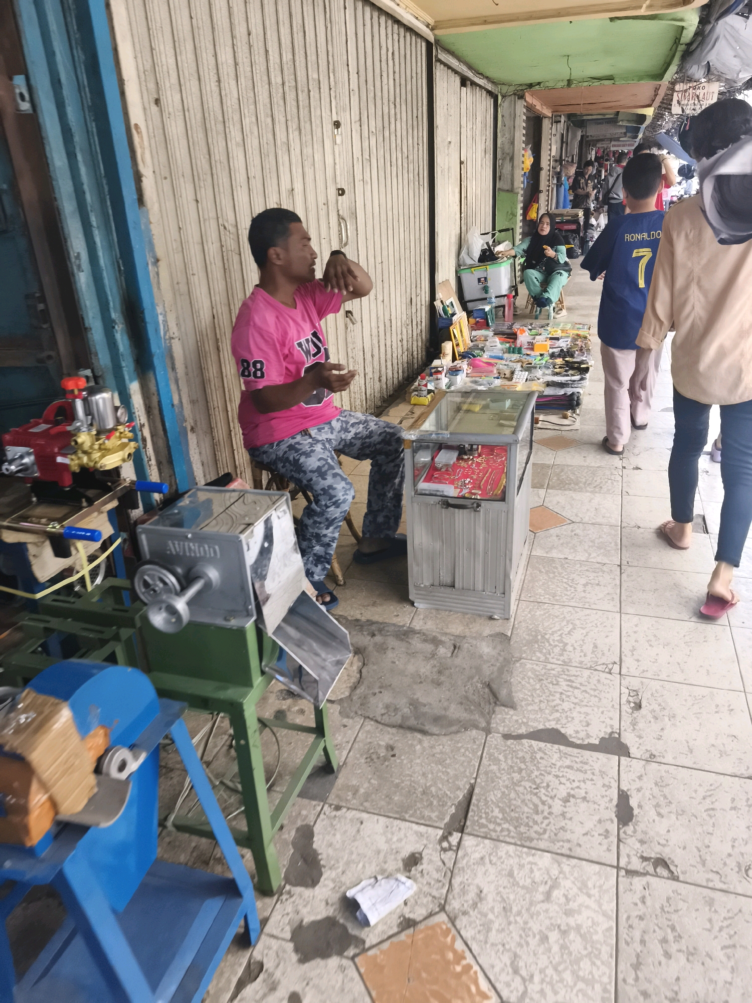 The image depicts a bustling street market in Kelurahan 16 Ilir, Kec. Ilir Timur I, Indonesia. A man wearing a pink t-shirt with the number "88" on it is seated on a stool, seemingly engaging in conversation or gesturing with his hands. In front of him, various items are displayed on a table and in a glass display case, including what appears to be jewelry and small electronic components. To his left, several mechanical devices are present, one a red and silver machine with blue handles, and another a silver and green contraption with a wheel, labeled "AVIMDO". In the background, other vendors and shoppers can be seen browsing the stalls, with a woman in a light blue hijab and green attire seated at a table, and a young person in a blue "RONALDO 7" jersey walking past. The market stalls are covered by a corrugated metal awning, and the ground is paved with square tiles. The overall impression is one of a vibrant, everyday marketplace.