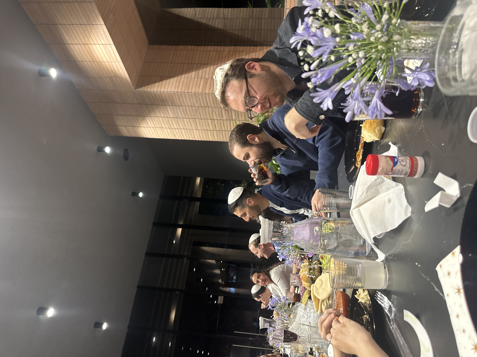 A long table is set for a meal. A group of people, many wearing kippahs, are seated along it. Plates of food, glasses, and vases of flowers are placed on the table. The setting appears to be indoors, possibly in a dining room. Outside, through a glass wall, lights can be seen. The location is inferred to be in Huixquilucan de Degollado, Mexico.
