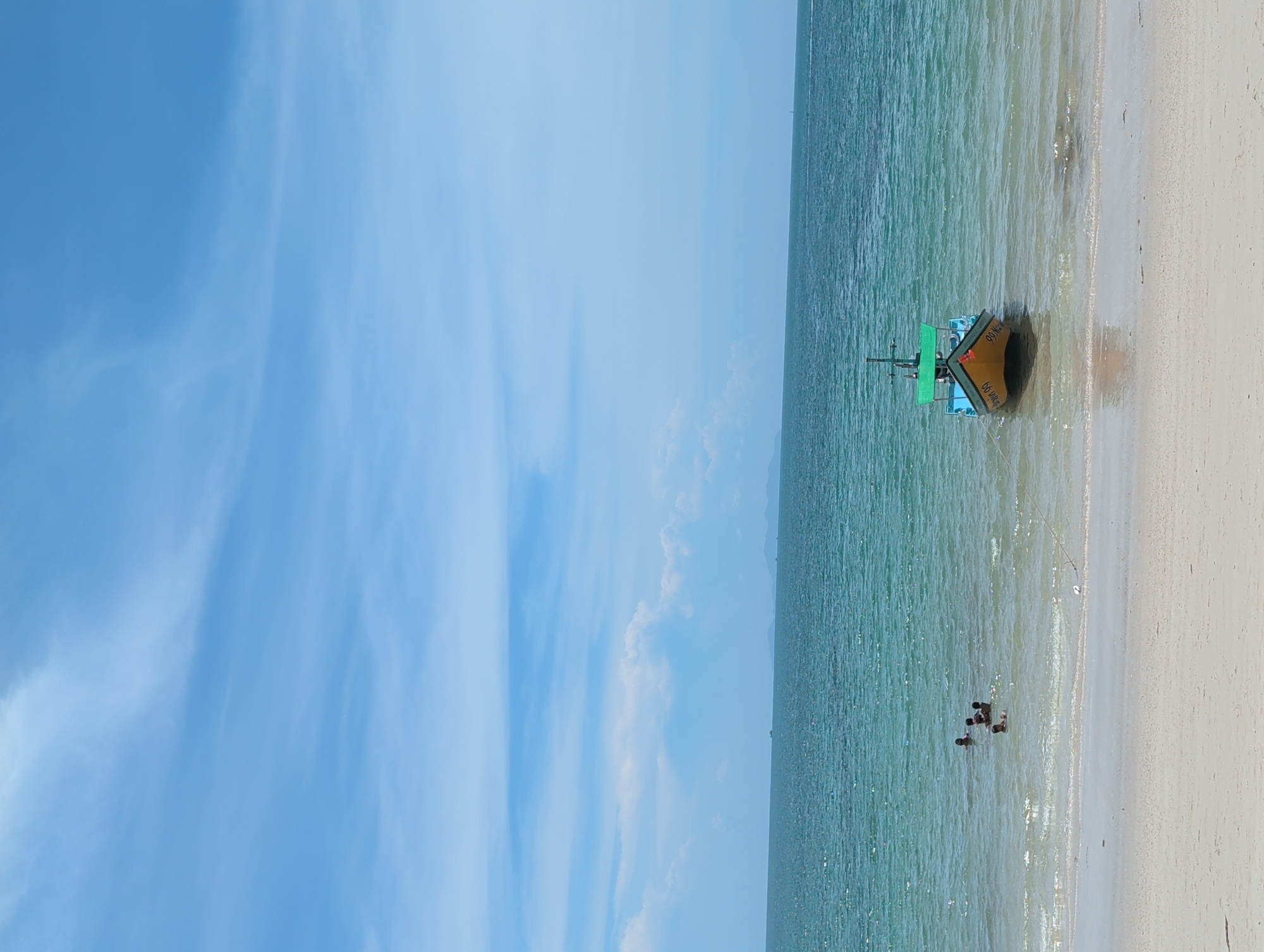 The image depicts a coastal scene in ตำบลเกาะพะงัน, Thailand. A fishing boat, predominantly yellow and green, rests partially on the sandy beach and extends into the water. Further out in the water, a small group of people are seen standing, possibly wading or swimming. The beach is sandy and light-colored. The water appears calm with a slight greenish hue. The sky is mostly blue with scattered white clouds.