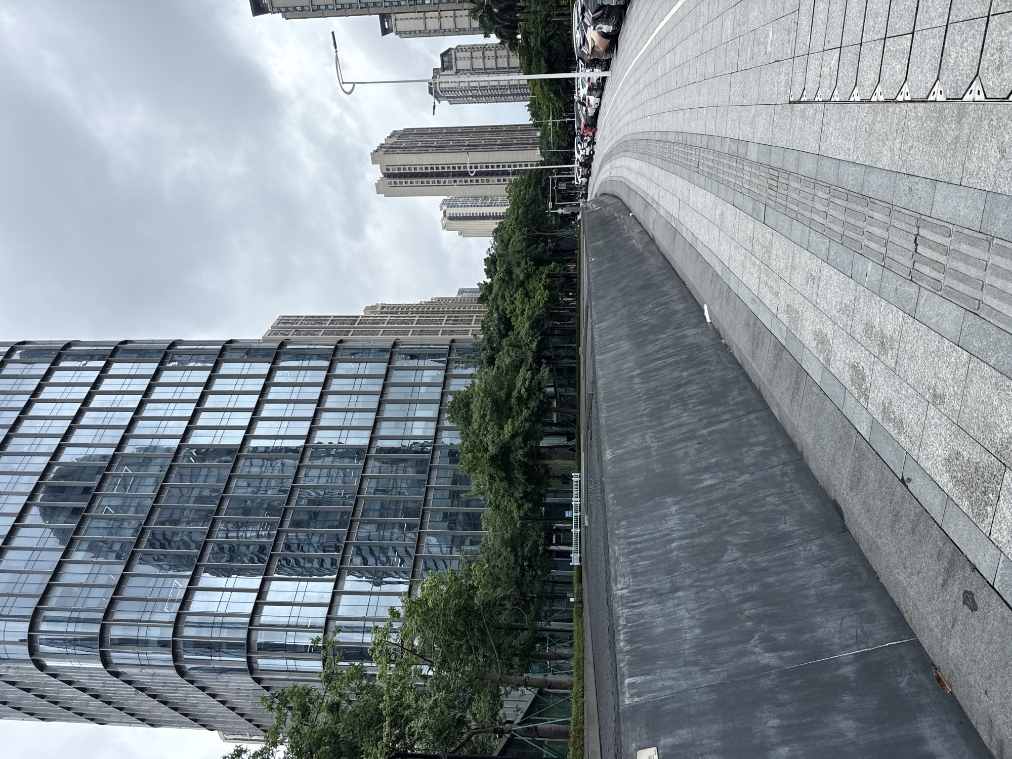 The image depicts a street scene in Haizhu District, China, characterized by modern architecture and urban infrastructure. In the foreground, a dark, sloped retaining wall runs along the left side of the frame, bordering a paved walkway. The walkway, composed of grey granite tiles with a tactile paving strip for visually impaired pedestrians, curves to the right.
To the left, a tall, glass-facaded building dominates the view. Its reflective surface mirrors the cloudy sky and the facades of other buildings. A line of lush green trees separates the retaining wall from the building's base, with some trees showing support structures.
Beyond the trees and the glass building, several other multi-story residential or office buildings of varying heights and architectural styles are visible in the background, stretching towards the horizon under an overcast sky. A street lamp with a curved arm stands prominently on the right side of the walkway.
A row of parked cars, including sedans and SUVs of various colors, are lined up along the right edge of the paved area, adjacent to the trees. Some of these vehicles are electric, indicated by visible charging ports or cables. A few motorcycles or scooters are also parked among the cars. The scene appears quiet, with no people immediately visible on the walkway or in the parked vehicles. The overall environment suggests a well-maintained urban public space.