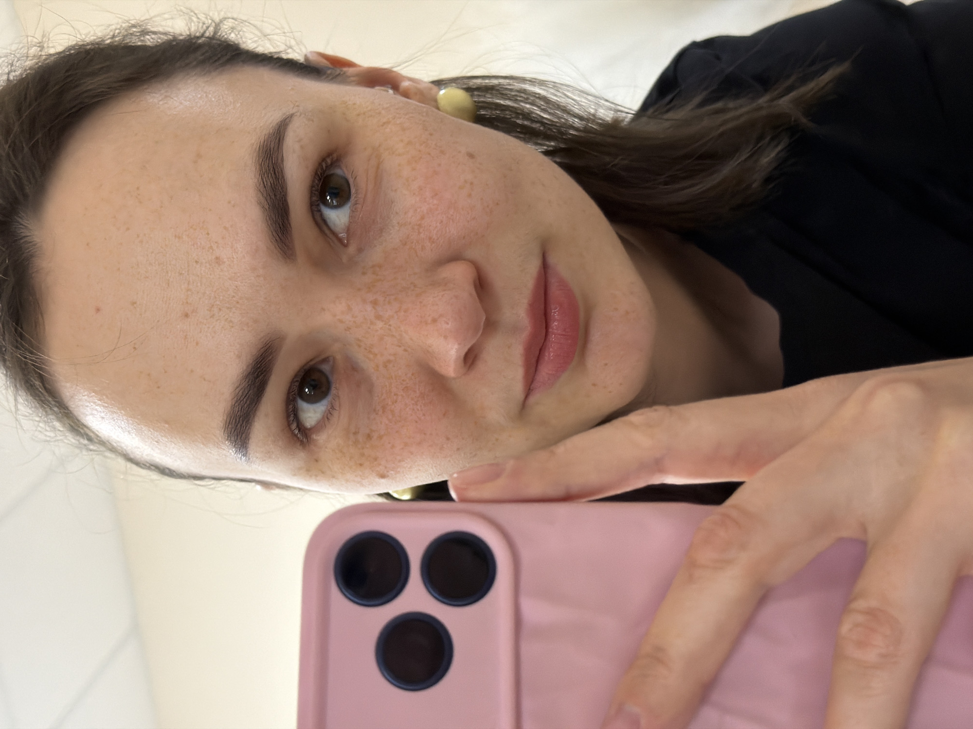A woman is holding a pink smartphone and taking a selfie. The woman has light skin, brown hair, and brown eyes. She has freckles on her face and is wearing a black top. The location is not identifiable as the city of Chaikovsky, Russia. The background is a light beige color.