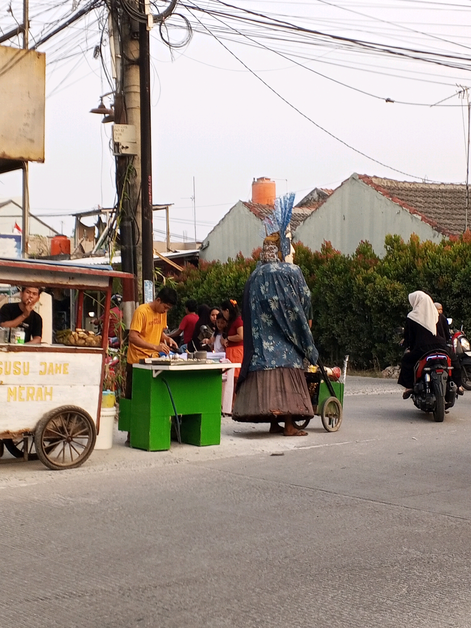 In Ciruas, Indonesia, on a street lined with houses and lush green bushes, a scene unfolds at dusk. A food cart, adorned with the words "SUSU JAHE MERAH" (red ginger milk), is positioned on the left side of the road. A man in a black shirt, likely a vendor, smokes a cigarette while leaning against the cart. Next to him, a vendor in a yellow shirt prepares food on a green table. A giant effigy, dressed in a blue floral patterned tunic and a brown skirt, stands near the food cart, its head adorned with blue feathers. To the right, a woman in a white hijab rides a motorcycle, while another motorbike is parked nearby. The sky is overcast, casting a soft, diffused light on the scene.