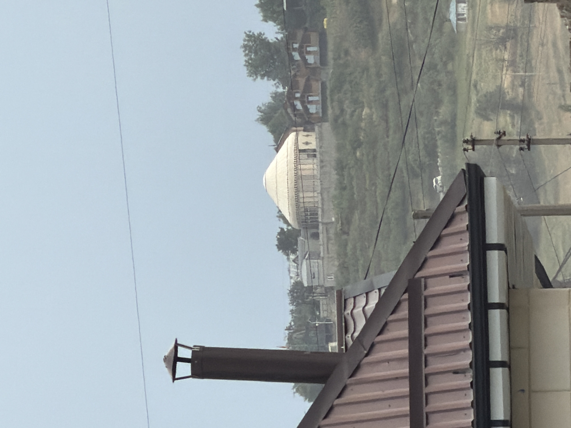 This media file captures an outdoor scene, likely in a rural or semi-rural setting. The most prominent object in the foreground is a portion of a building's roof, constructed with reddish-brown corrugated metal. Attached to this roof is a dark-colored chimney pipe, which extends upwards and appears to be made of metal. The roofline is angled, suggesting it's part of a house or another structure.
In the mid-ground, a large, dome-shaped structure is visible, appearing to be white or a very light color, possibly a yurt or a geodesic dome. This structure is surrounded by greenery and other smaller buildings that resemble houses. Some of these houses have light-colored walls and darker roofs, with visible windows. The background consists of a sloping, vegetated hill or embankment, with more trees and some distant structures partially obscured by a hazy or overcast sky. Several power lines are strung across the scene, supported by utility poles.
The time of day appears to be daytime, but the sky is overcast, creating a diffused light and a slightly muted color palette. The weather seems to be hazy or possibly humid. There are no people clearly visible in the media file. The overall impression is of a residential area nestled amongst natural landscape, with some modern elements like the power lines and the dome structure. There is no visible text in the image.